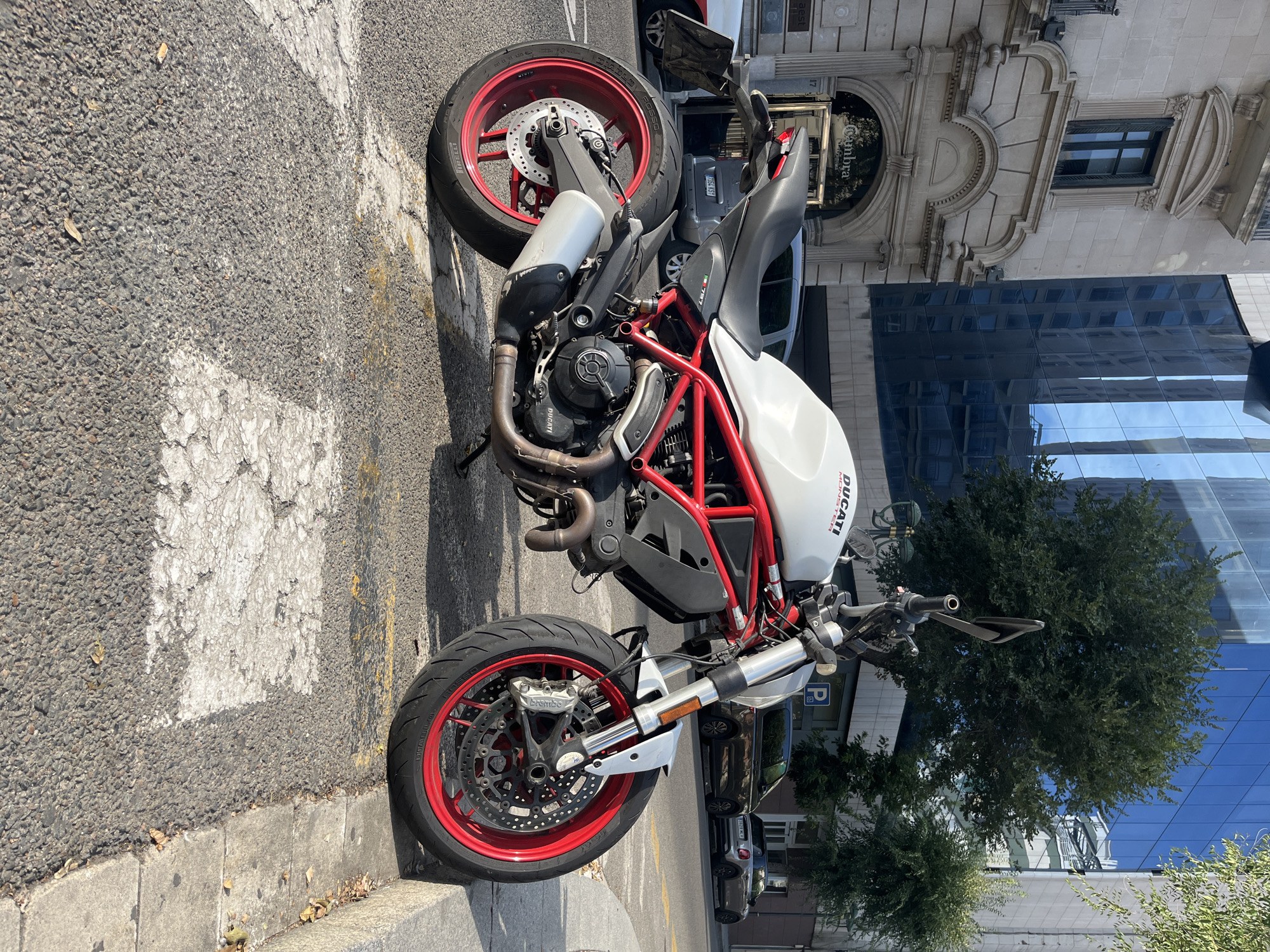 The image captures a Ducati Monster motorcycle parked on a pedestrian crossing on a sunny day in Tarragona, Spain. The motorcycle, primarily white with a red frame, is positioned facing right, with its front wheel slightly turned towards the sidewalk. The background features a mix of old and modern architecture, including a building with ornate stonework and arched entrances, and a contemporary building with a blue glass facade reflecting the sky and surrounding trees. Cars are visible parked on the street behind the motorcycle. The pedestrian crossing stripes are faded and cracked, indicating wear. The overall scene depicts an urban streetscape with a focus on the parked motorcycle.