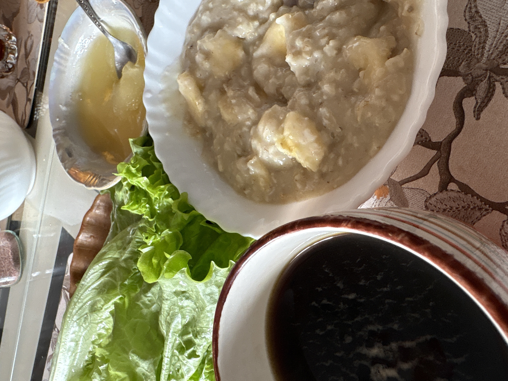A table displays a white bowl filled with oatmeal and sliced bananas, accompanied by a small dish of what appears to be stewed apples, and a bowl of dark liquid, likely tea or coffee, alongside a serving of green lettuce. The scene takes place indoors, possibly in a dining area with a floral-patterned tablecloth. There are no people visible. It is impossible to confirm the location in the city of Ulyanovsk, Russia based on this image alone.