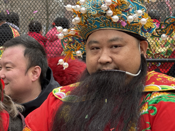 A man in elaborate traditional attire is the central focus of the image, captured from the chest up. He is of East Asian descent, likely middle-aged, with dark hair and a serious, direct gaze towards the viewer.
He wears a vibrant red robe, richly embroidered with intricate patterns in gold, green, and possibly other colors, visible on his shoulders and collar. On his head rests a large, ornate headdress, predominantly gold and blue, adorned with numerous white spherical pearls that appear to be part of an intricate design, some dangling from wires or branches on the hat.
A long, full black beard, which appears to be artificial, extends down from his chin. A thin, white wire, possibly part of a microphone or headset, runs from near his right ear across his cheek and below his mouth, featuring a coiled black section at its center.
To the left of the main subject, partially visible, is another man of East Asian descent, smiling and looking off to the left. He has shorter dark hair and is wearing a dark jacket. In the background, a chain-link fence separates the subjects from a blurred crowd of people, some of whom are wearing red clothing, suggesting a public gathering or celebration. The overall setting appears to be outdoors during the daytime.
The elaborate costume, particularly the headdress and artificial beard, suggests the main subject is portraying a specific character or figure, possibly for a cultural performance, parade, or festival, such as a Chinese New Year celebration, given the style of dress. No discernible text is visible in the image.