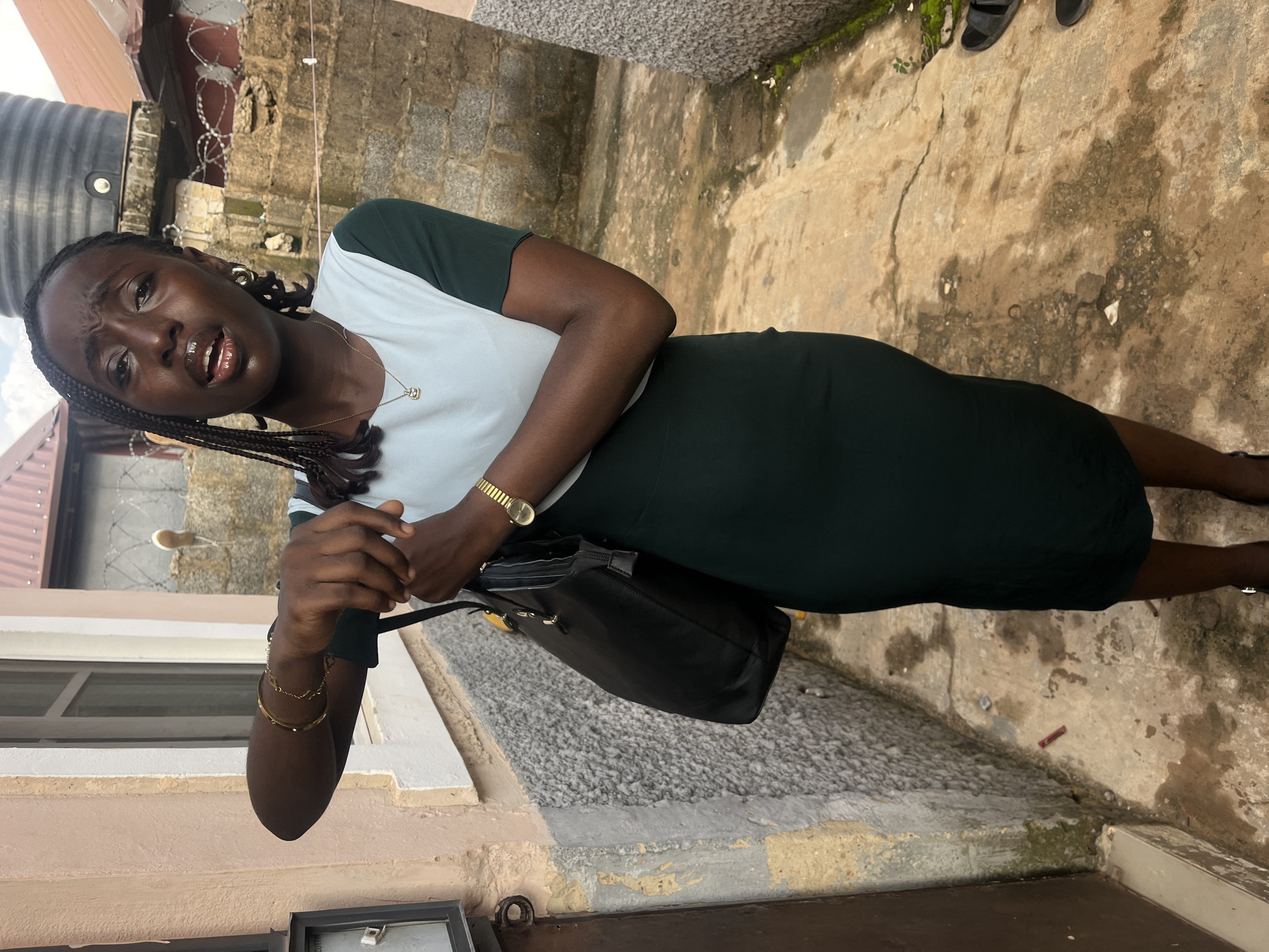A young Black woman, with dark skin and braided hair, stands in what appears to be an alleyway with concrete walls and a gritty, stained ground. She is wearing a two-toned dress, light blue on top and dark green on the bottom, with short sleeves. She has on gold bangles and a delicate gold necklace with a small pendant. A black handbag hangs from her left shoulder, and she is holding its strap with her right hand. Her left arm is bent at the elbow, and her left hand is positioned near her face, almost as if she's about to touch it. Her mouth is open, suggesting she is speaking or surprised.
The background reveals a rough-textured stone wall, a section of a corrugated metal roof, and a large black water tank. Barbed wire is visible along the top of the wall. The ground is a mix of cracked concrete and dirt, with some patches of moss and scattered debris, including a small red object. The overall impression is a somewhat weathered, possibly residential, urban environment. Based on the architectural style of the buildings, the water tank, and the overall atmosphere, this scene is likely located in a neighborhood in Abuja, Nigeria.