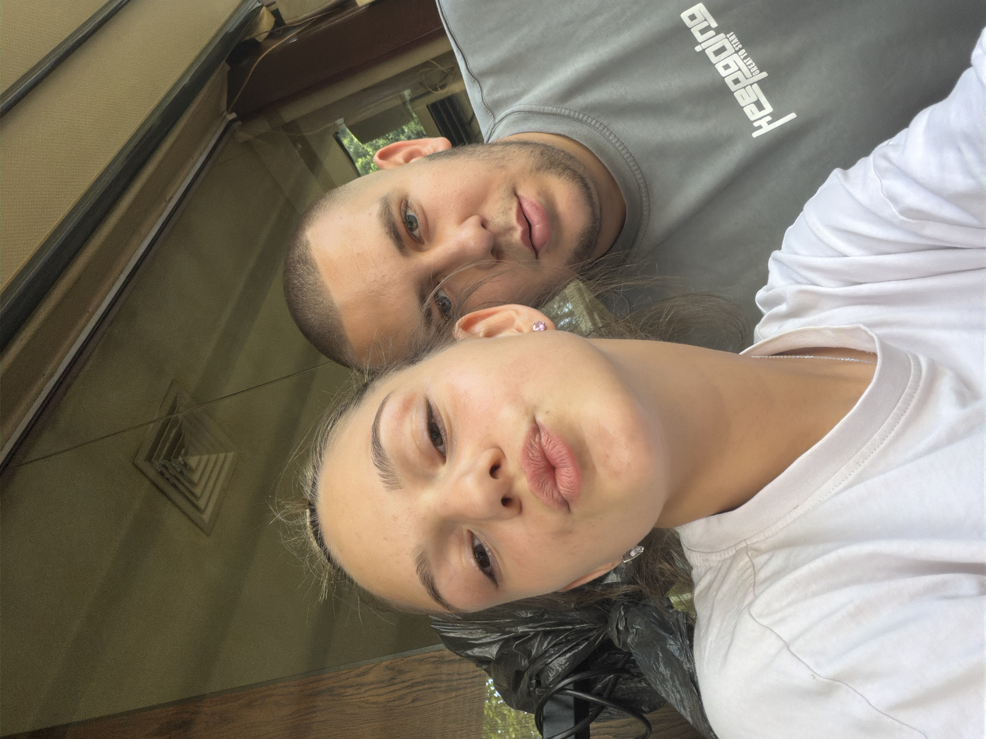 Two people are in the image. A man with short hair and a beard is wearing a gray shirt. A woman with long hair is wearing a white shirt. They appear to be taking a selfie. The location is Troyan, Bulgaria. The environment appears to be outdoors near a building.