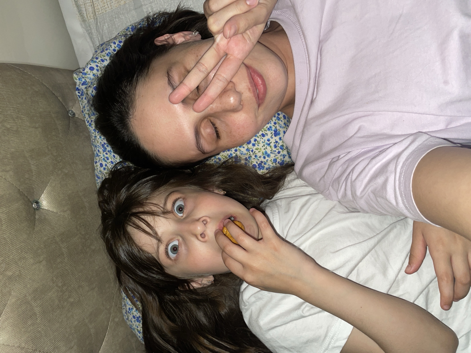 Two individuals, likely a mother and daughter, are lying on a bed or cushioned surface. The woman is older, with dark hair and fair skin, wearing a light purple shirt and making a peace sign over one eye. The girl, with brown hair and wide blue eyes, is holding a round object in her mouth, possibly a cookie or piece of food. The scene is indoors, with a patterned pillow under the woman's head. There is a tufted or patterned surface visible on the left, which could be a couch or headboard. Location is unknown; there is no indication of Izhevsk, Russia.