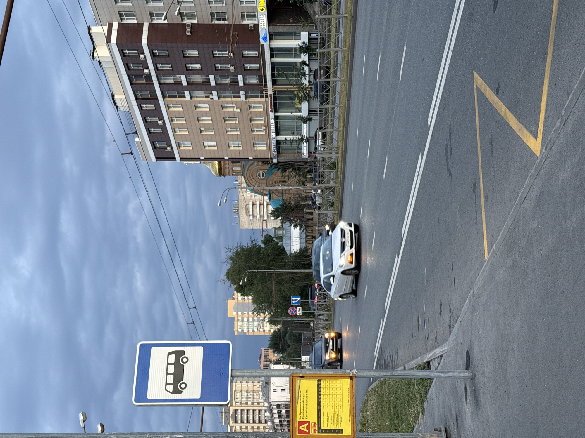 The image captures a street scene in Kazan, Russia, with a bus stop sign prominently displayed in the foreground. The sign, featuring a black bus silhouette on a blue background, is positioned next to a yellow informational board with Russian text, likely detailing bus routes and schedules. A silver car is driving on the wet asphalt road, with another dark-colored vehicle behind it. In the background, a modern residential building with multiple stories and a gold-domed church stand against a cloudy sky. The overall environment suggests a city street during daylight, possibly after rain.