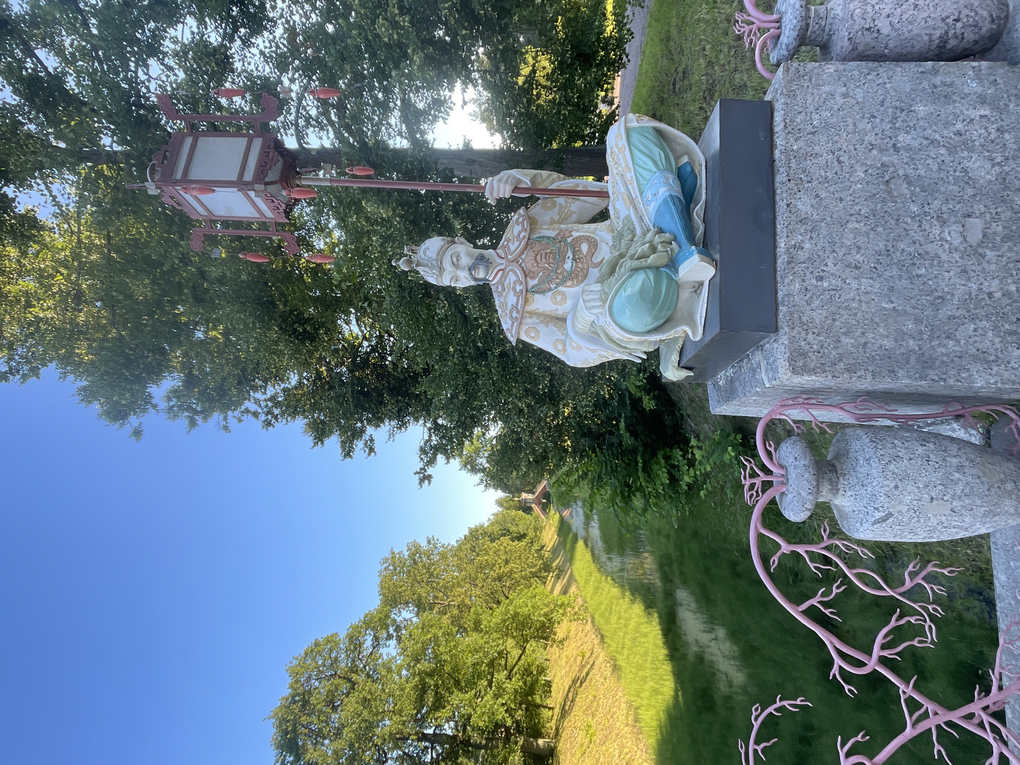 A statue of a seated Asian man holding a lantern-like object is positioned on a stone pedestal in the foreground. The man is dressed in ornate, colorful clothing, including a headdress, robes with dragon motifs, and blue leggings. Behind the statue, a canal with lush green grass banks is visible, leading to a small arched bridge in the distance. The area is surrounded by mature trees with green foliage, and the sky is a clear, bright blue. The scene appears to be set in a park or garden. This is likely the Chinese Bridge in Catherine Park, located in the city of Pushkin, Russia.