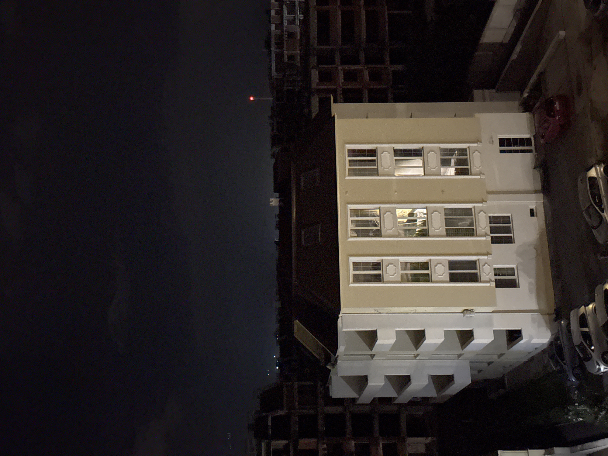 The image captures an urban night scene in Plovdiv, Bulgaria, with two prominent buildings under a dark sky. The central subject is a multi-story building, likely an apartment or office block, featuring a light beige facade on its upper floors and a white lower section with distinct architectural elements resembling deep, inverted pyramid-shaped alcoves. Numerous rectangular windows with white frames are visible, some reflecting interior lights, and a few are adorned with white louvered shutters.
Behind this structure, a taller, dark building stands, appearing to be an unfinished construction site. Its skeletal framework of concrete or steel beams is discernible against the night, and a small, red warning light or beacon glows near its top.
To the lower right, a portion of a street or parking area is visible, with several cars parked. A bright red sedan and a silver hatchback are clearly identifiable. The overall atmosphere is quiet, characteristic of an urban environment after dark. No people or specific activities are depicted, and no visible text can be seen in the frame.