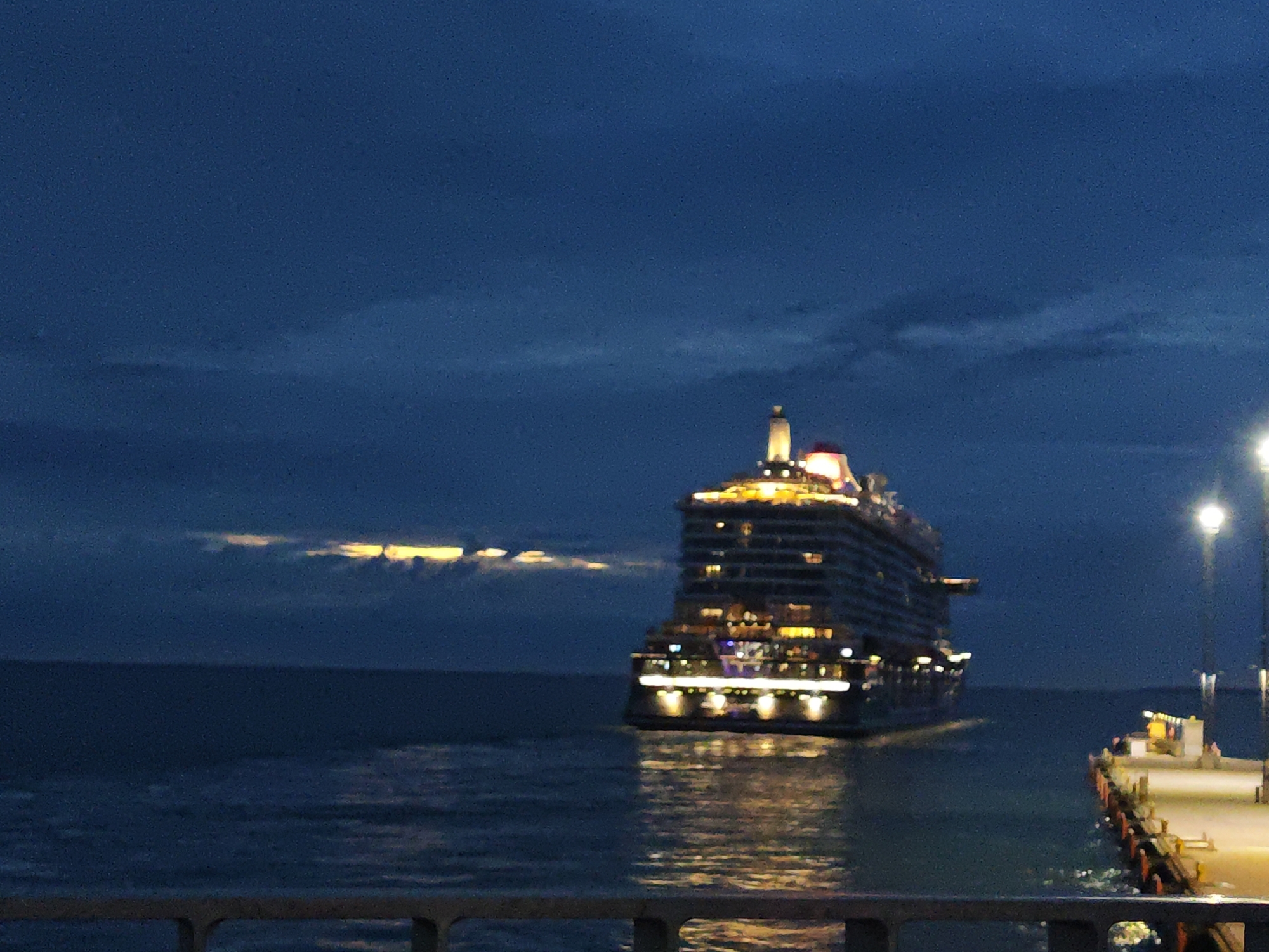 This media file depicts a large cruise ship docked at a pier in Tallinn, Estonia during twilight. The ship is illuminated with numerous lights, highlighting its size and multiple decks. The water around the ship reflects the lights, creating a shimmering effect. The sky is a deep blue, with some scattered clouds that are catching the last rays of sunlight, appearing as a band of yellow and orange on the horizon. On the right side of the image, a pier is visible with lampposts and some small structures. The overall mood is serene and peaceful, capturing the transition from day to night. There is no visible text in the image.