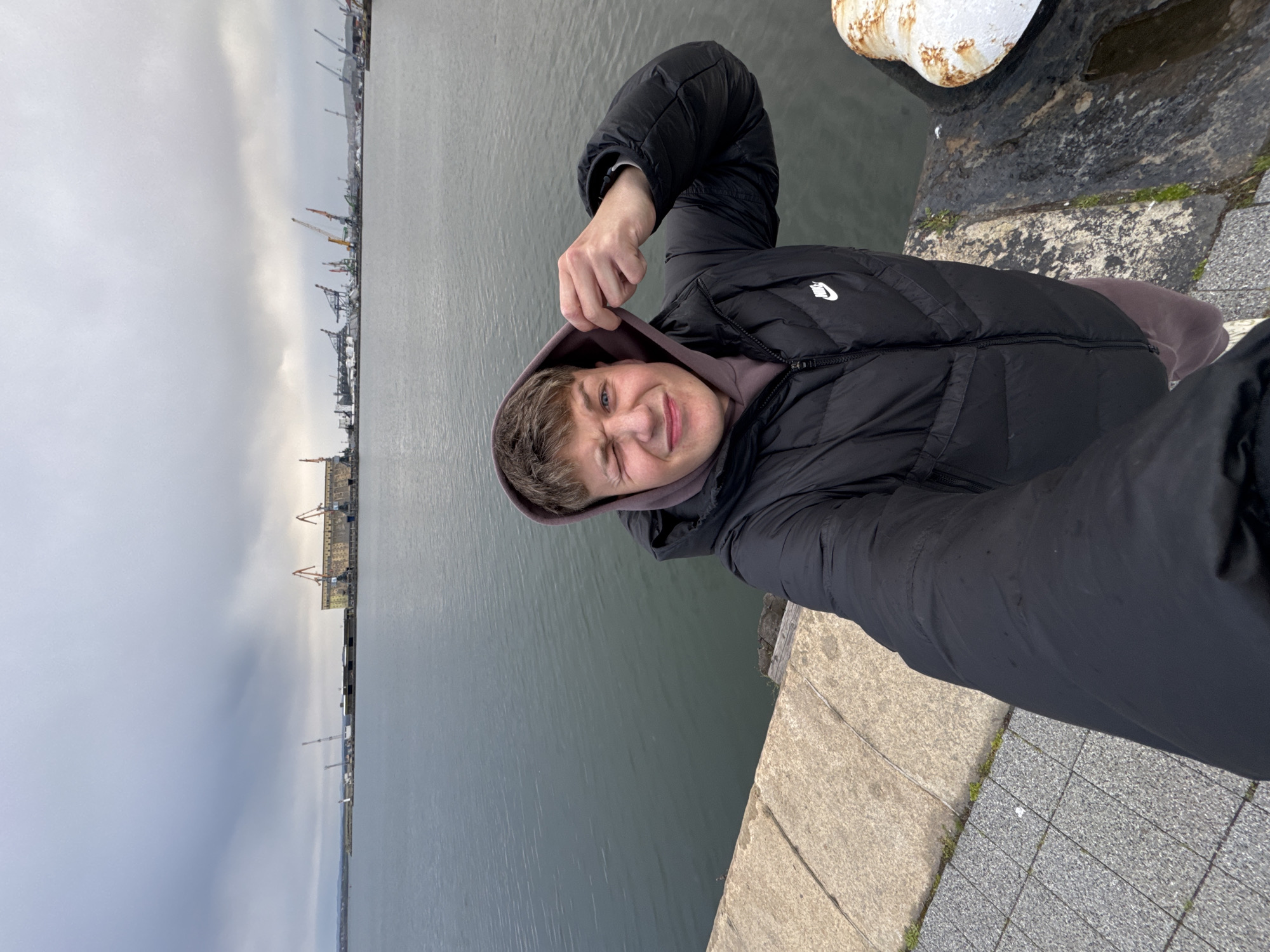 A young man stands on a concrete pier along the waterfront, looking at the camera. He is wearing a black puffer jacket over a brown hooded sweatshirt. He is holding the hood of his sweatshirt with his left hand. The pier is made of concrete slabs, with small patches of grass growing in the cracks. Next to the pier is a white bollard with rust stains. Behind the man is a body of water, and in the distance, there is a harbor with cranes and buildings. The sky is overcast. The photograph appears to be taken in Burgas, Bulgaria, based on the presence of port infrastructure that is characteristic of that city.