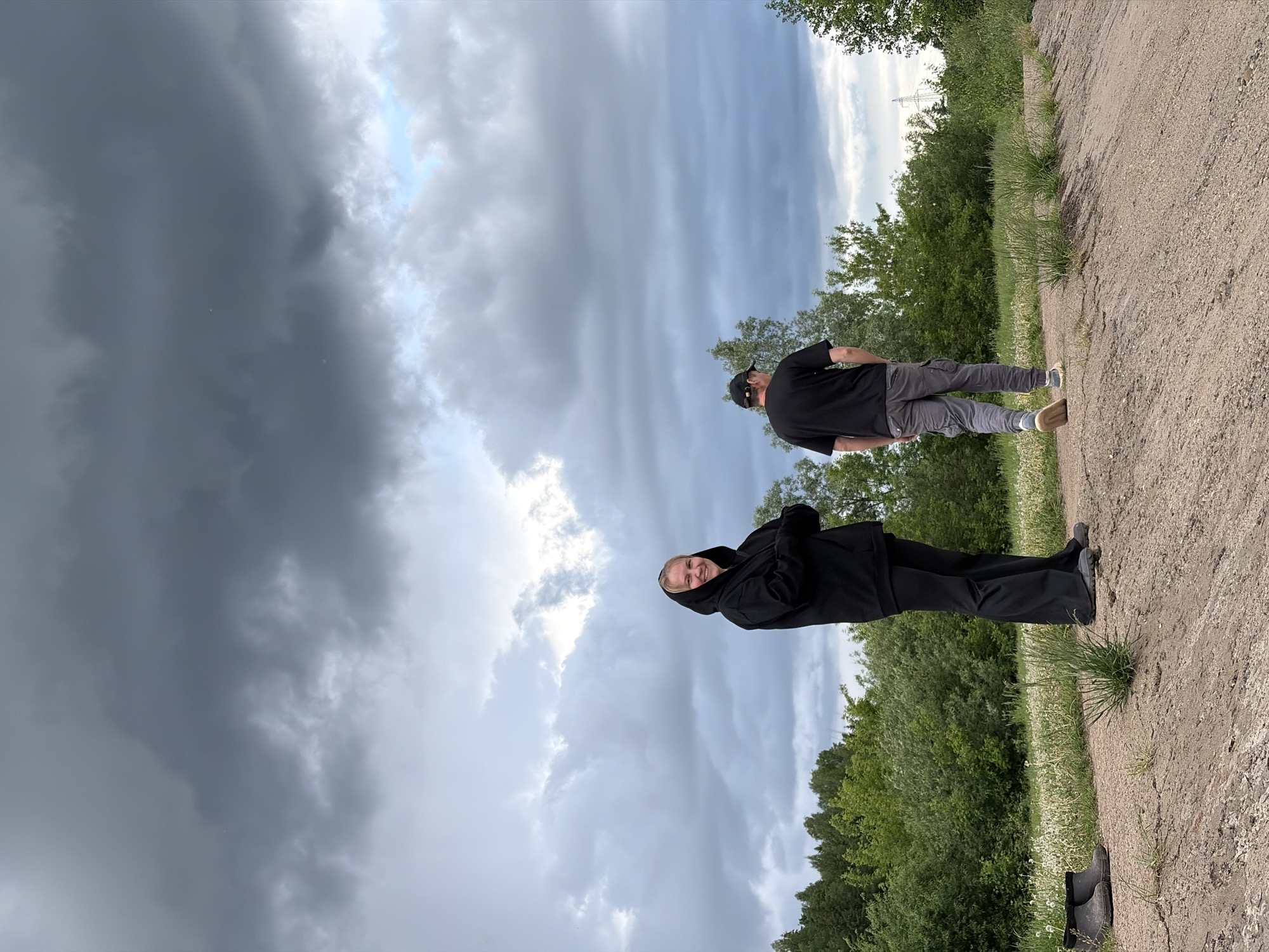 The image shows two people on a dirt road with green vegetation on either side and a cloudy sky above.
The person closer to the camera is wearing a black hooded jacket and black pants. They are smiling and looking at the camera.
The person further away is wearing a black shirt, grey pants, a black baseball cap, and sunglasses. They are facing away from the camera.
In the background, there is a power line visible among the trees.
Based on information available, it is not possible to definitively identify the specific location as Nizhnii Vishur, Russia, without further evidence.