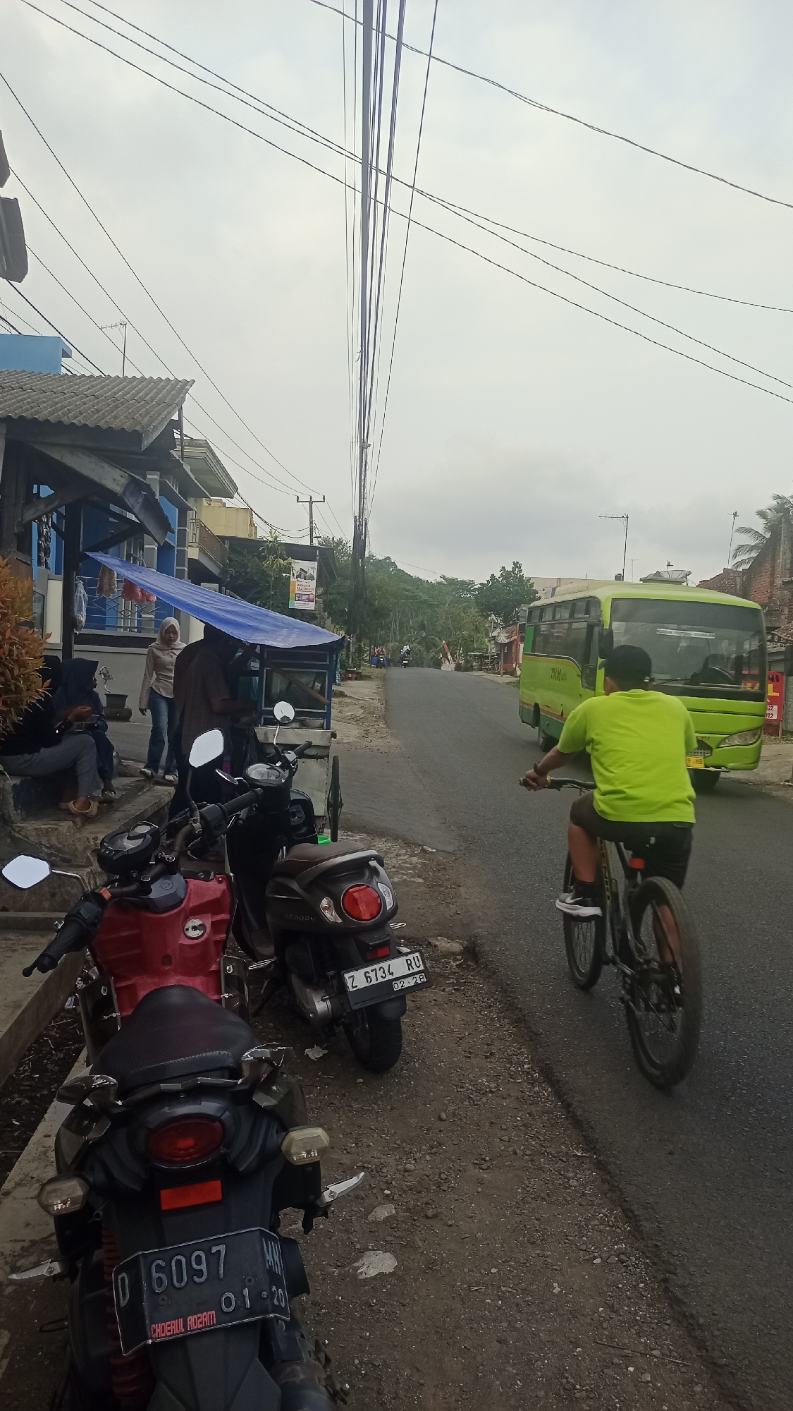 The image depicts a street scene in Karyamandala, Indonesia, likely in a rural or semi-urban area. In the foreground, there are several motorcycles parked on a dirt and gravel shoulder adjacent to an asphalt road. A red motorcycle is on the left, followed by a dark-colored scooter, and then a darker motorcycle with a license plate that reads "D 6097". On the road, a person wearing a bright green t-shirt and a black cap is riding a bicycle, moving away from the viewer. To the right, a green bus is driving down the road in the same direction as the cyclist. On the left side of the road, there is a structure with a blue tarp providing shade over a small food stall or vendor area. Several people are visible near this stall; two women are seated on steps, and two individuals are standing near the stall. Overhead, numerous electrical wires crisscross the sky. The environment appears to be overcast, with a cloudy sky.