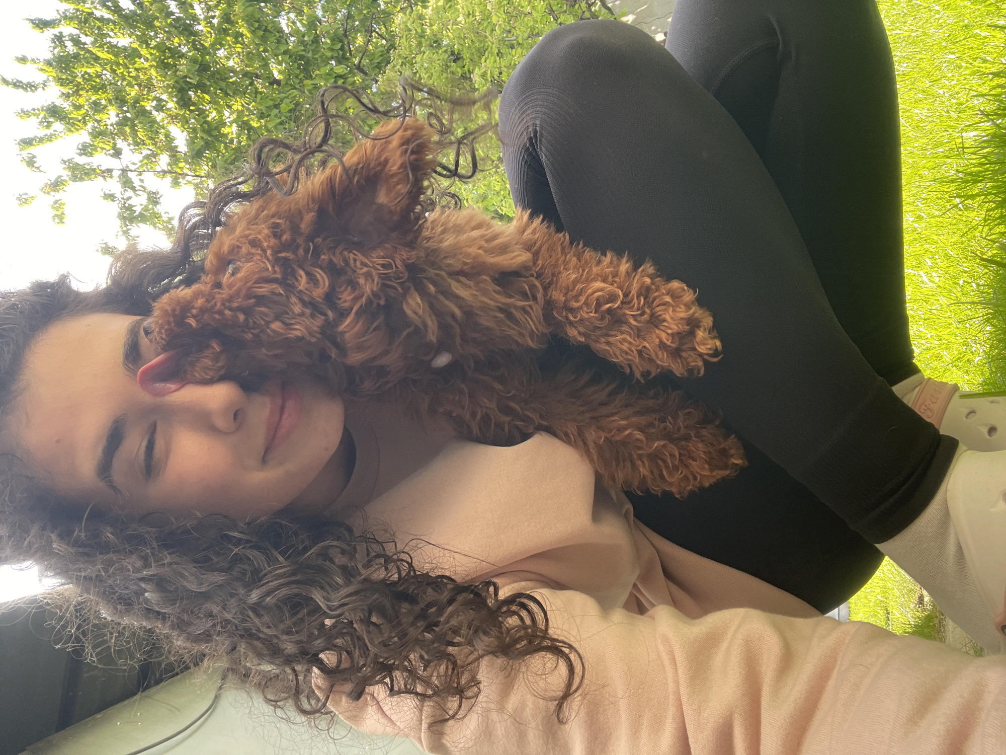 A young woman with curly brown hair is outdoors, possibly in a park, with a small brown curly-haired dog resting on her chest. She is lying down, angled towards the viewer, wearing a light pink long-sleeved shirt and black leggings. The dog appears to be licking her face. Sunlight filters through trees in the background. The scene is located on a grassy area, which suggests a park or green space. There is no visible indication that it is the city of Sofia, Bulgaria.