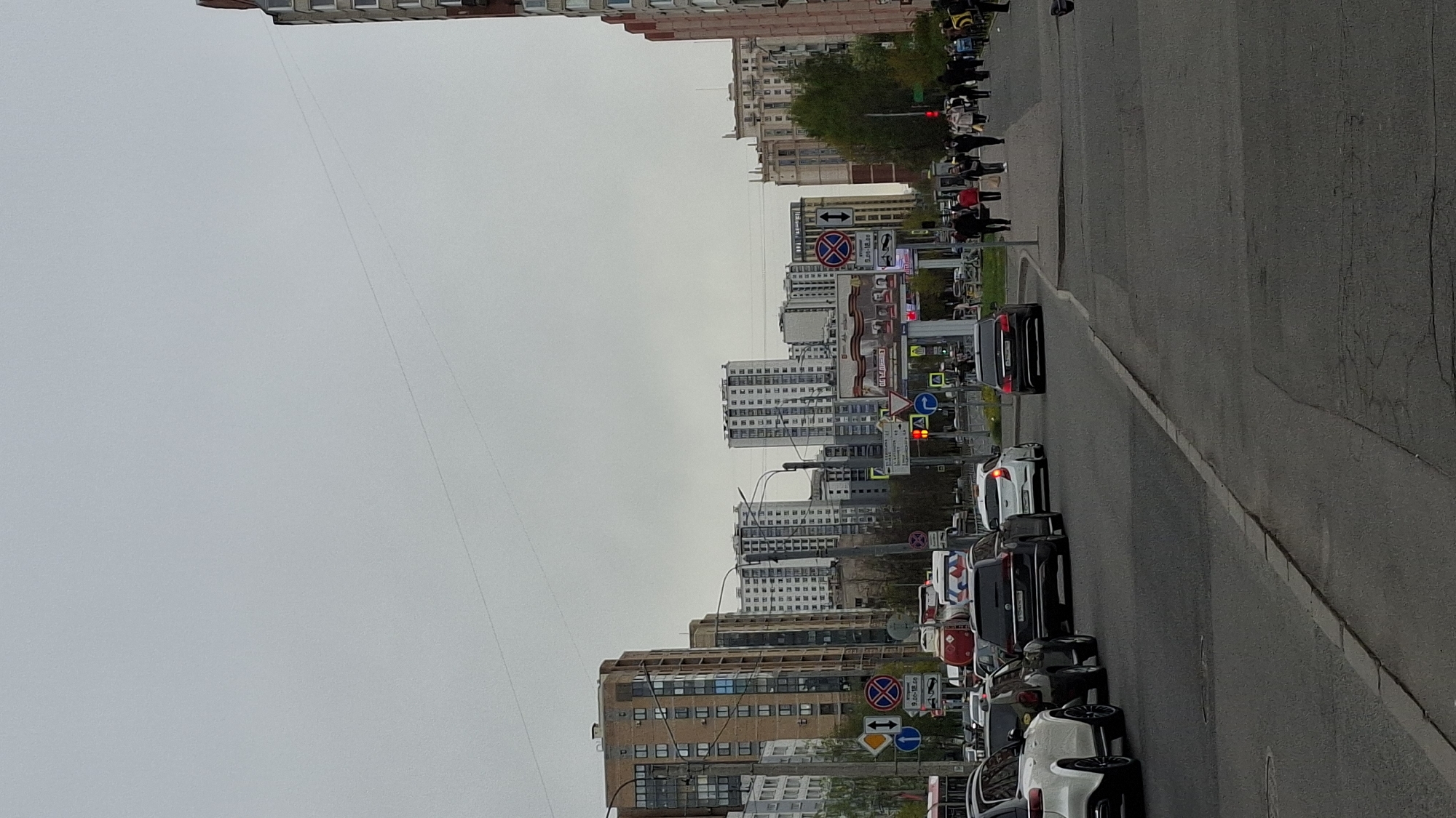 The image depicts a street scene in Saint Petersburg, Russia, characterized by dense urban development and vehicular traffic. The road runs along the right, with a line of parked cars and moving vehicles. Various types of cars and vans are observed, mixed with traffic signs indicating parking restrictions and mandatory directions. A pedestrian crossing with people waiting to cross can also be seen. The urban environment includes high-rise residential buildings with apartment units. The sky is overcast, suggesting a cloudy day. Electrical wiring is visible stretched across the sky between buildings.