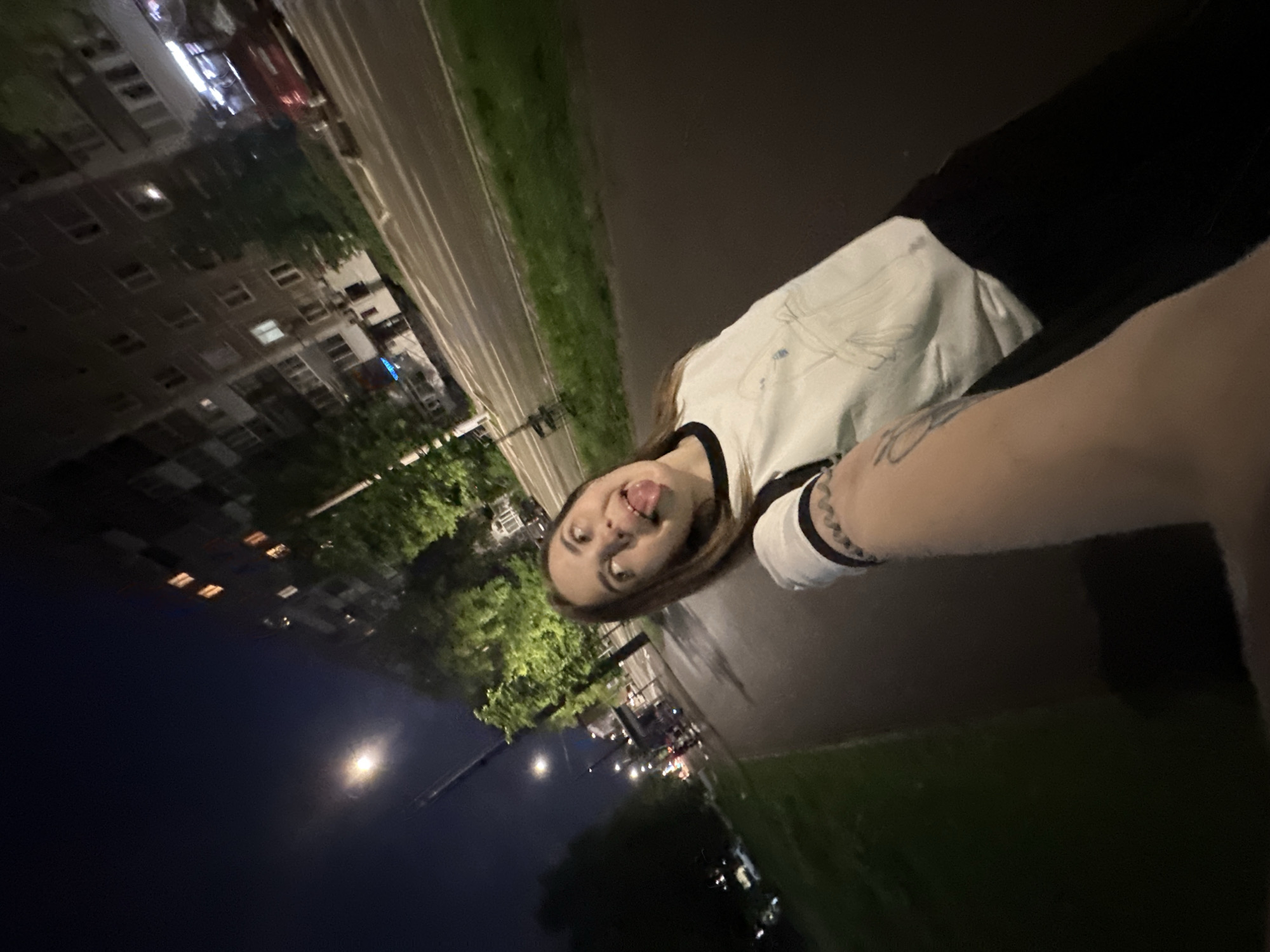 A woman with light brown hair and fair skin, wearing a white and black long-sleeved shirt, is posing for a selfie at night in what appears to be Perm, Russia. She is sticking her tongue out and has a tattoo on her left arm. The photo is taken at an angle, with city buildings and trees visible in the background on the left. A street and green spaces run through the scene, illuminated by streetlights. The overall lighting is low, contributing to a nighttime ambiance.