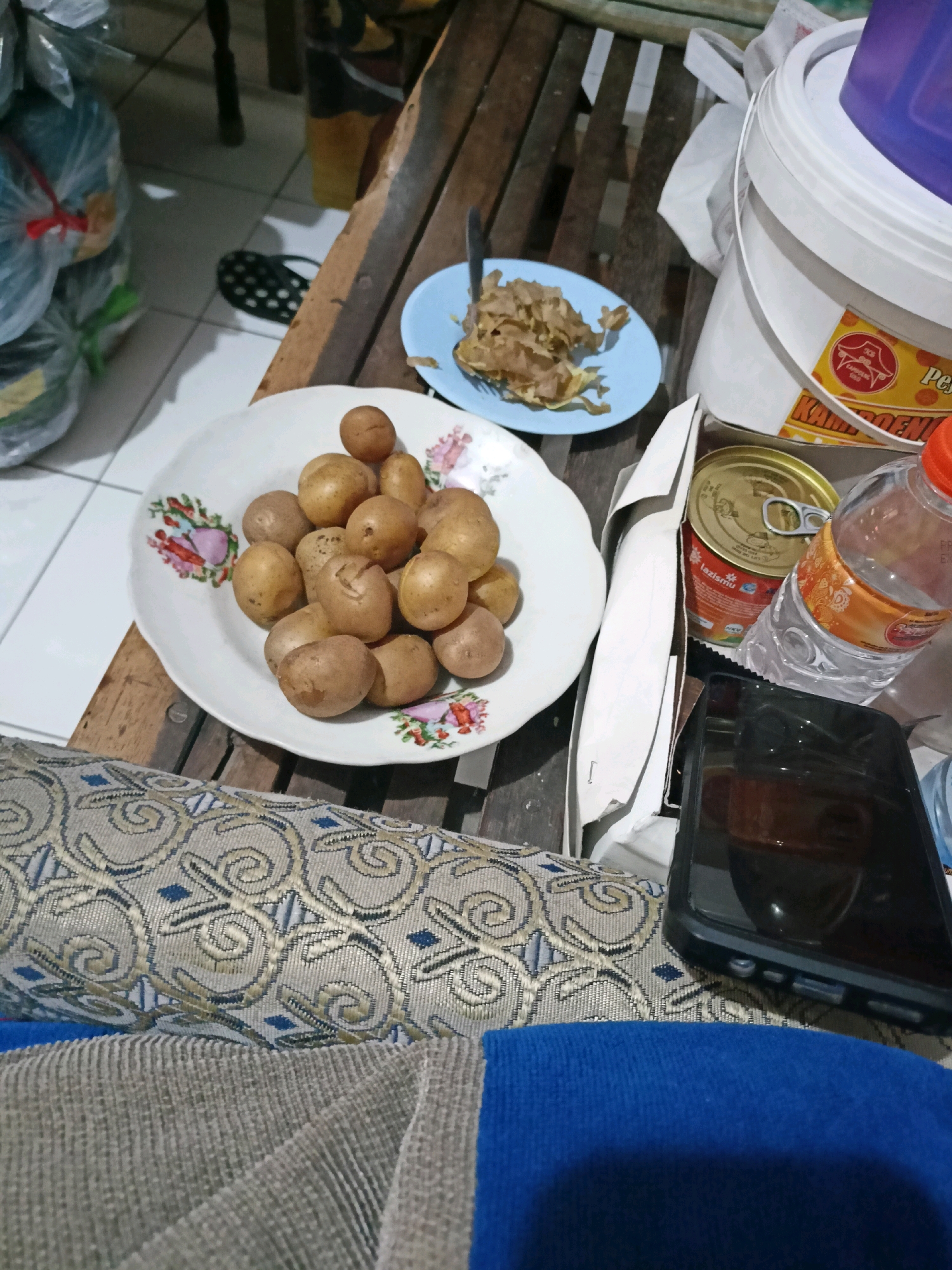 The image depicts a close-up view of a wooden table laden with food items and a mobile phone. On the table, a white plate holds a pile of small, unpeeled potatoes. Next to it, a small blue plate contains a portion of what appears to be fried food. To the right, a large white bucket with an orange label is visible, and a partially obscured clear plastic bottle with an orange cap sits next to it. A metallic can with a pull-tab is also present. In the foreground, a black mobile phone with a dark case rests on a patterned fabric. The background shows a tiled floor and some large, plastic-wrapped items. The scene appears to be indoors, possibly in a home in the city of Payaman, Indonesia, given the context.