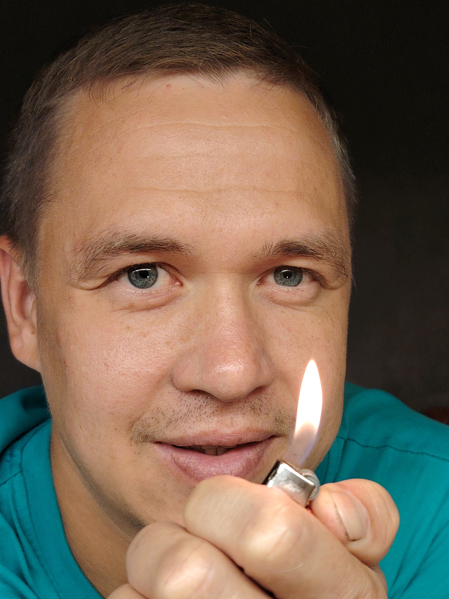 A man with light brown hair and blue eyes is shown in a close-up portrait, holding a lit lighter near his face. He is wearing a teal t-shirt. The background is dark, suggesting an indoor setting. The man's expression is neutral. The image does not provide any information about a specific location in Chaikovsky, Russia.