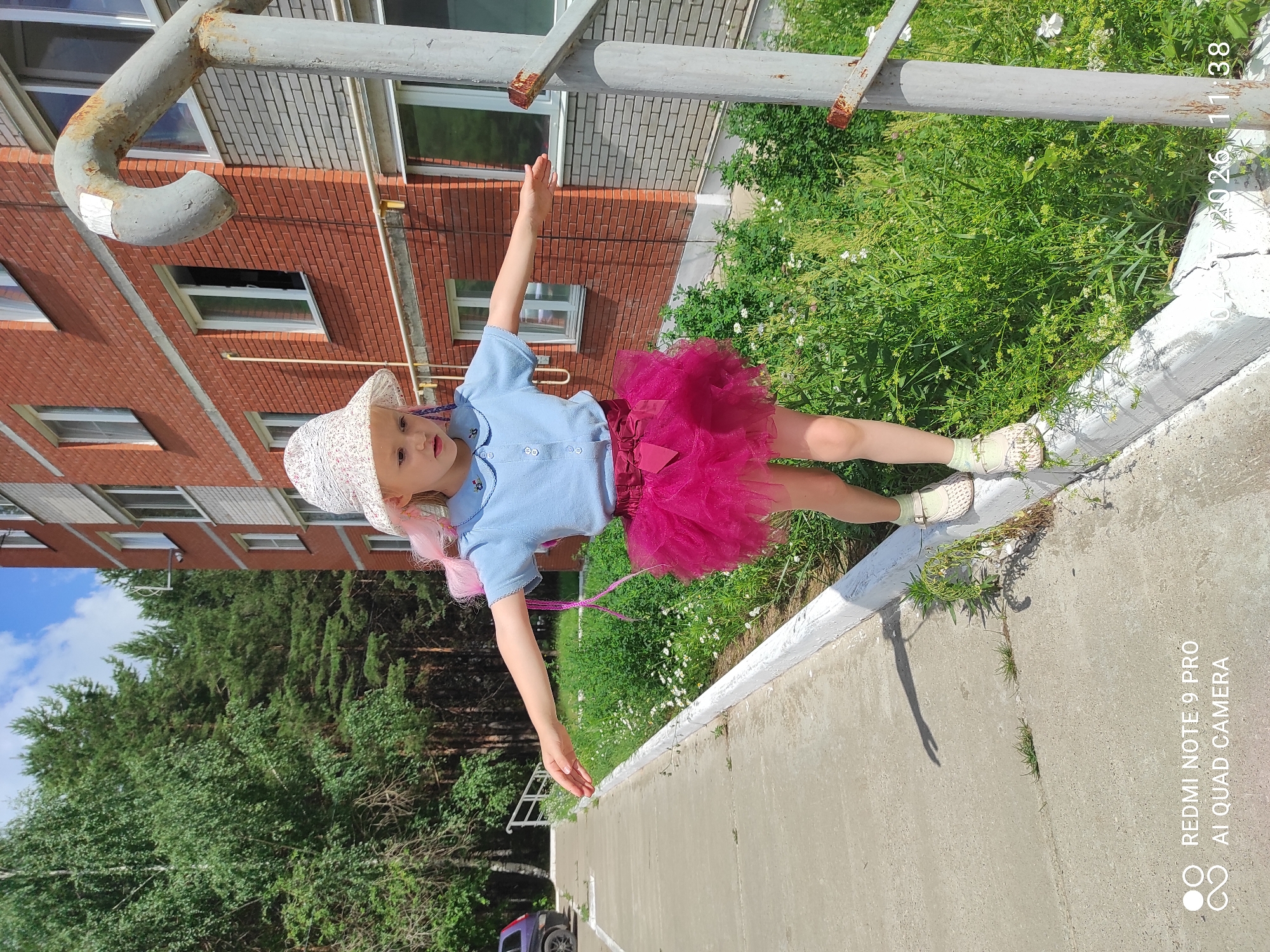 A young girl with blonde hair, adorned with pink streaks, wearing a light blue collared shirt, a pink tutu, a white patterned sun hat, and beige clogs, is captured in a full-body shot. She is balancing on a concrete curb lined with white paint, her arms extended sideways for equilibrium. The background features a multi-story brick apartment building, a dense green forest, and a gray metal railing. The lighting suggests a bright, sunny day. The image metadata indicates it was taken in Chaikovsky, Russia, on April 7, 2026, at 11:38 AM.