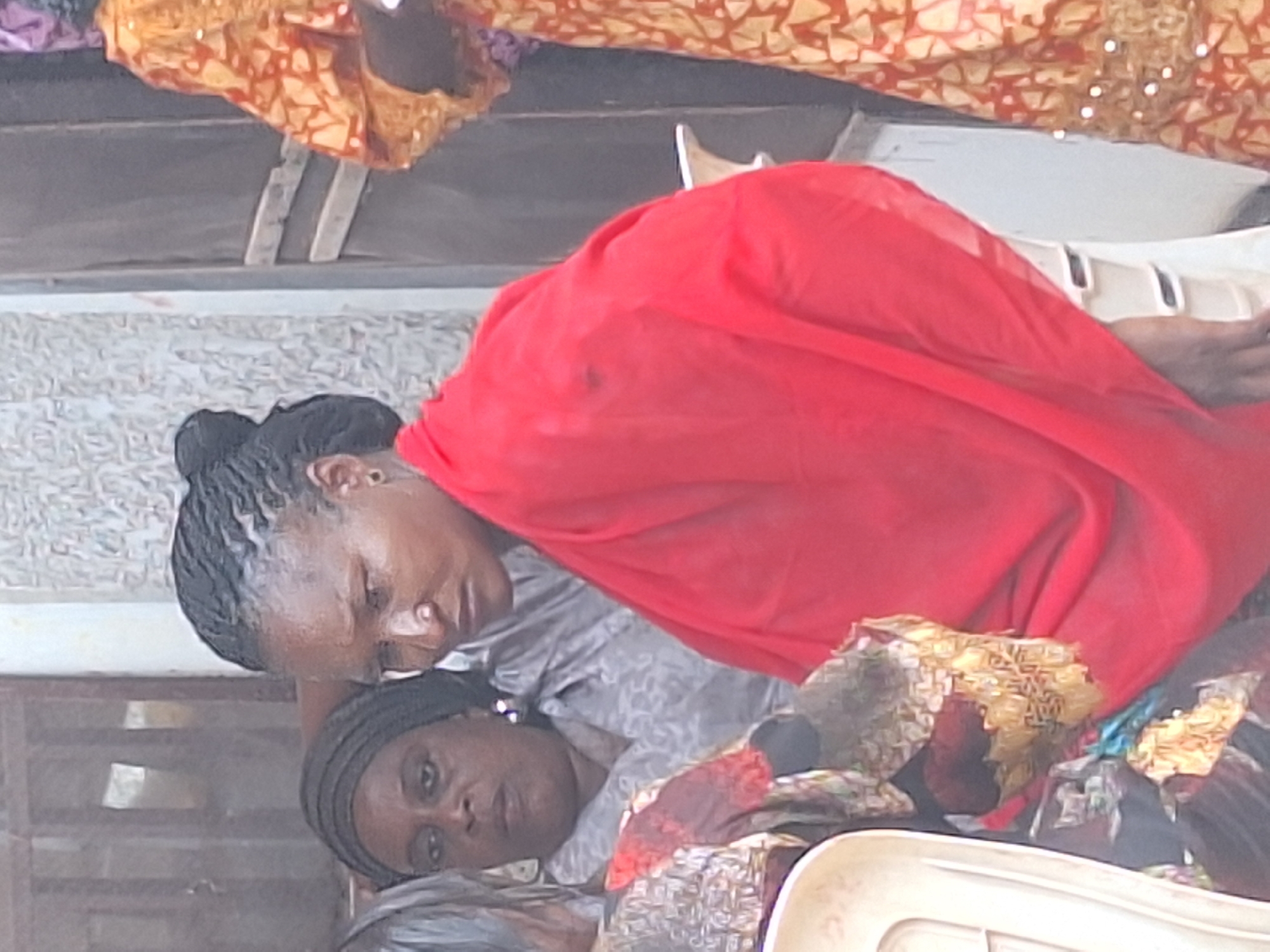 Two women are in Sabon Tasha, Nigeria. One woman is lying down, while the other is sitting next to her. The woman sitting next to her has a red scarf draped over her shoulder.