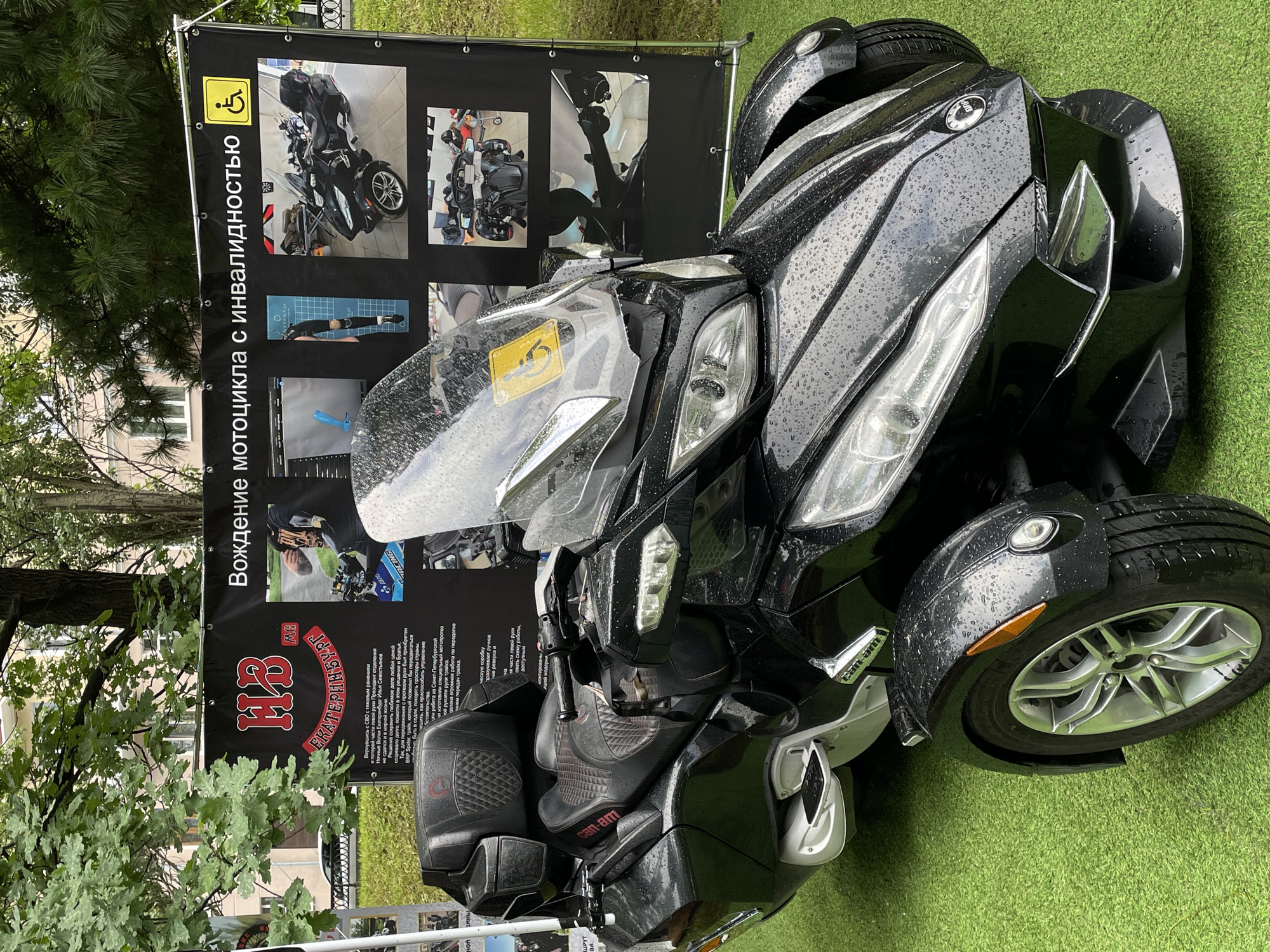 A black Can-Am Spyder three-wheeled motorcycle, heavily coated with small water droplets, is prominently displayed on artificial green turf in an outdoor setting in Yekaterinburg, Russia. Behind the vehicle, a large black banner serves as a backdrop, detailing "Motorcycle Riding with Disability" (Вождение мотоцикла с инвалидностью) and featuring a yellow wheelchair accessibility symbol in the top right corner.
The banner also displays the logo "НВ МС ЕКАТЕРИНБУРГ" (HV MC YEKATERINBURG) in red and white, identifying it as an initiative by the Night Wolves MC motorcycle club's Yekaterinburg chapter. Text on the banner explains the adaptation of motorcycles for individuals with disabilities, specifically mentioning a veteran named Ilya Sevostyanov, President of the Night Wolves Yekaterinburg chapter, who lost parts of his left arm after returning from the Special Military Operation (СВО). The text highlights the use of a BRP Spyder for mobility and adaptations for single-hand control. Multiple images on the banner illustrate prosthetic limbs, CAD designs for adaptations, close-ups of adapted handlebars, and various photographs of the Can-Am Spyder model. One image shows a person's tattooed forearm gripping an adapted handlebar.
The motorcycle itself features a large clear windshield, a black two-person seat with a quilted pattern, and integrated rear storage. A yellow square sticker with the international symbol of access (wheelchair) is affixed to the windshield. Silver-colored footrests are visible on both sides.
The surrounding environment indicates a park-like or urban green space. Lush green trees, including both deciduous and coniferous types, are visible in the background, along with a multi-story building featuring windows. A glimpse of a white car is partially visible to the far left. The ground in the immediate vicinity of the display is artificial turf, contrasting with the natural foliage further back. Overcast lighting conditions are suggested by the uniform light and the presence of raindrops on the motorcycle.