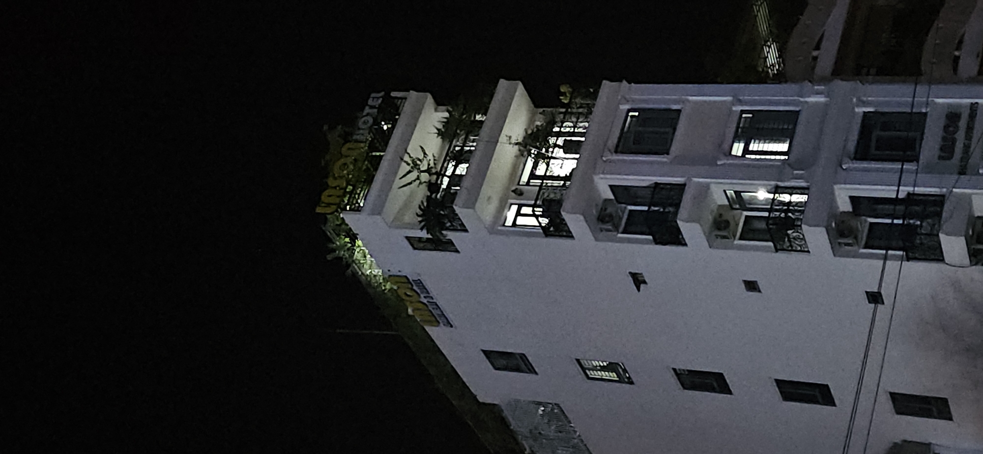 The image shows a hotel building at night in Bắc Nha Trang, Vietnam. The building is white and several stories high, with balconies on each level. There are plants on some of the balconies. Windows are visible, some with lights on inside. The hotel's name, "Insun Hotel," is written on the side of the building. The background is dark, indicating nighttime. There are utility wires in the shot.