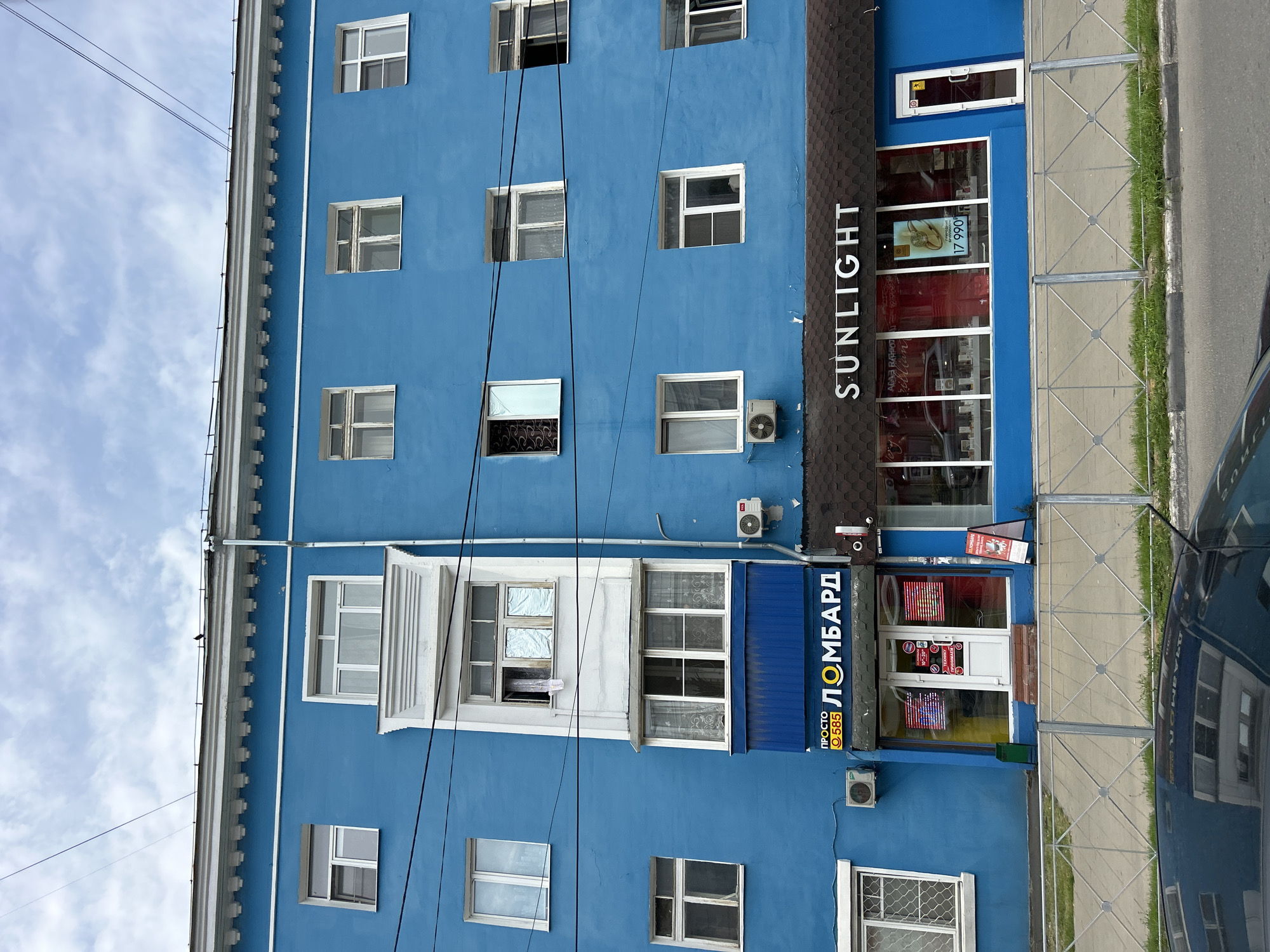 The image shows a blue building in the city of Lyubertsy, Russia. The building has several windows and a balcony. On the ground floor, there are two shops: a pawn shop with a sign that says "PROSTO 585 LOMBARDE" and a jewelry store with a sign that says "SUNLIGHT". A metal fence runs along the sidewalk in front of the shops, and a car is parked on the street beyond the fence. The sky is cloudy.