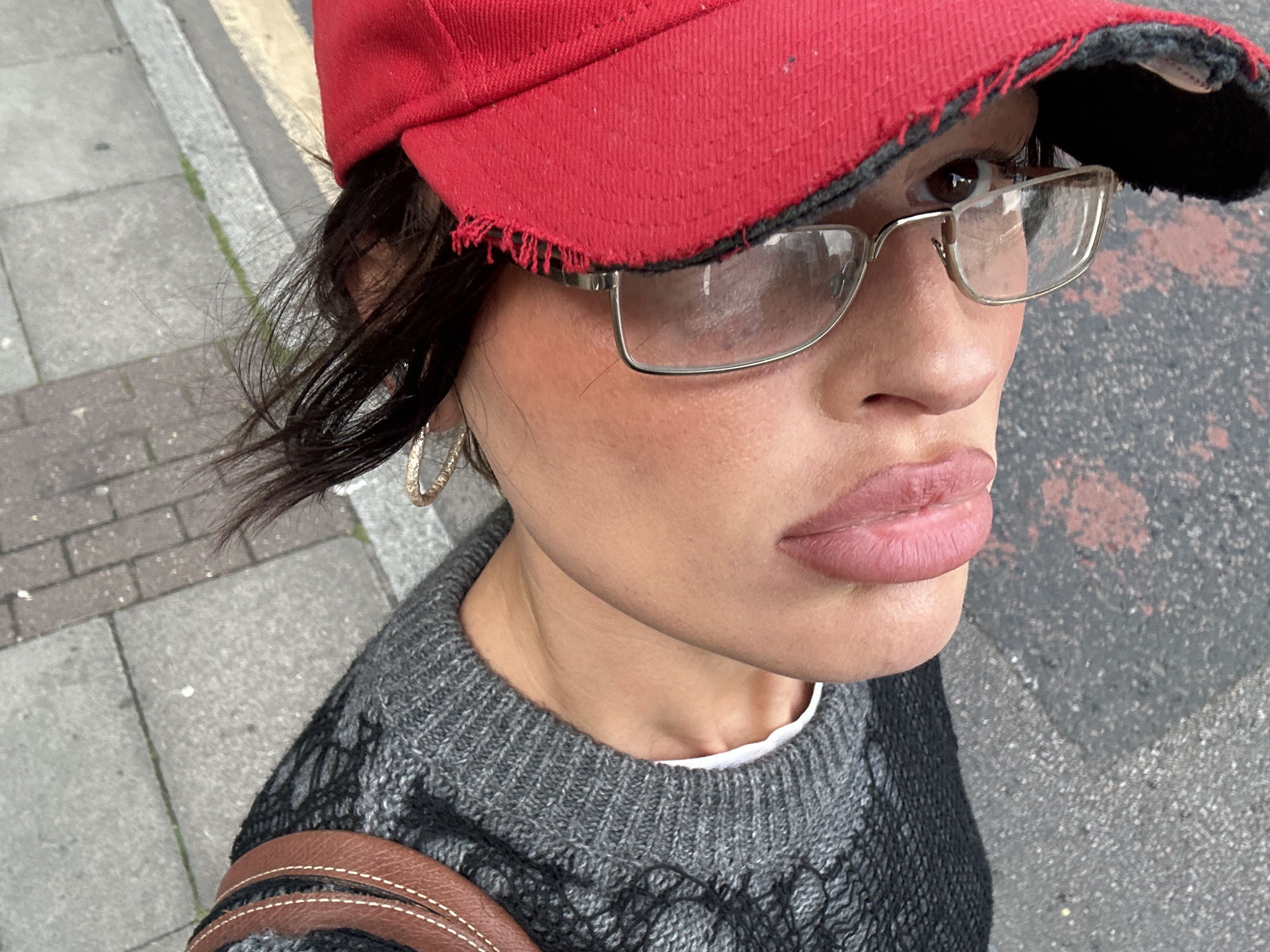 The image is a close-up selfie taken outdoors, likely on a city street. The subject is a woman wearing a red baseball cap with a frayed brim and silver-framed glasses. She has hoop earrings and wears a gray sweater with black knit detailing. Part of a brown leather bag strap is visible on her shoulder. The background shows a sidewalk made of gray rectangular paving stones and a street with a mottled gray surface, suggesting it might be asphalt or concrete. Based on the style of paving stones and general appearance of the background, the location is potentially within a city in the United Kingdom, with the city of London being a plausible option.