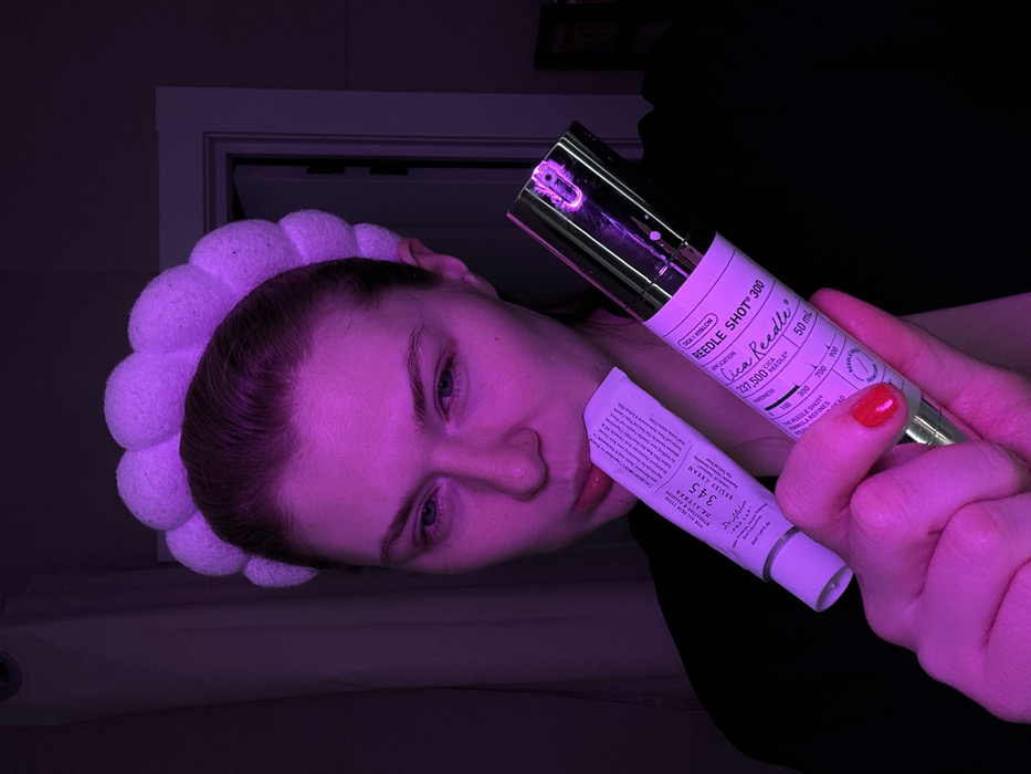 A person with a white, puffy headband is holding two skincare products under purple light. The product closer to the person's face is a white tube of "345 REALFIT RELIEF CREAM". Behind it, a hand with red nail polish holds a silver tube labeled "REEDLE SHOT 300". The label indicates it contains "Cica Reedle" with an application of "237,500 NEEDLES" in a 50ml container.