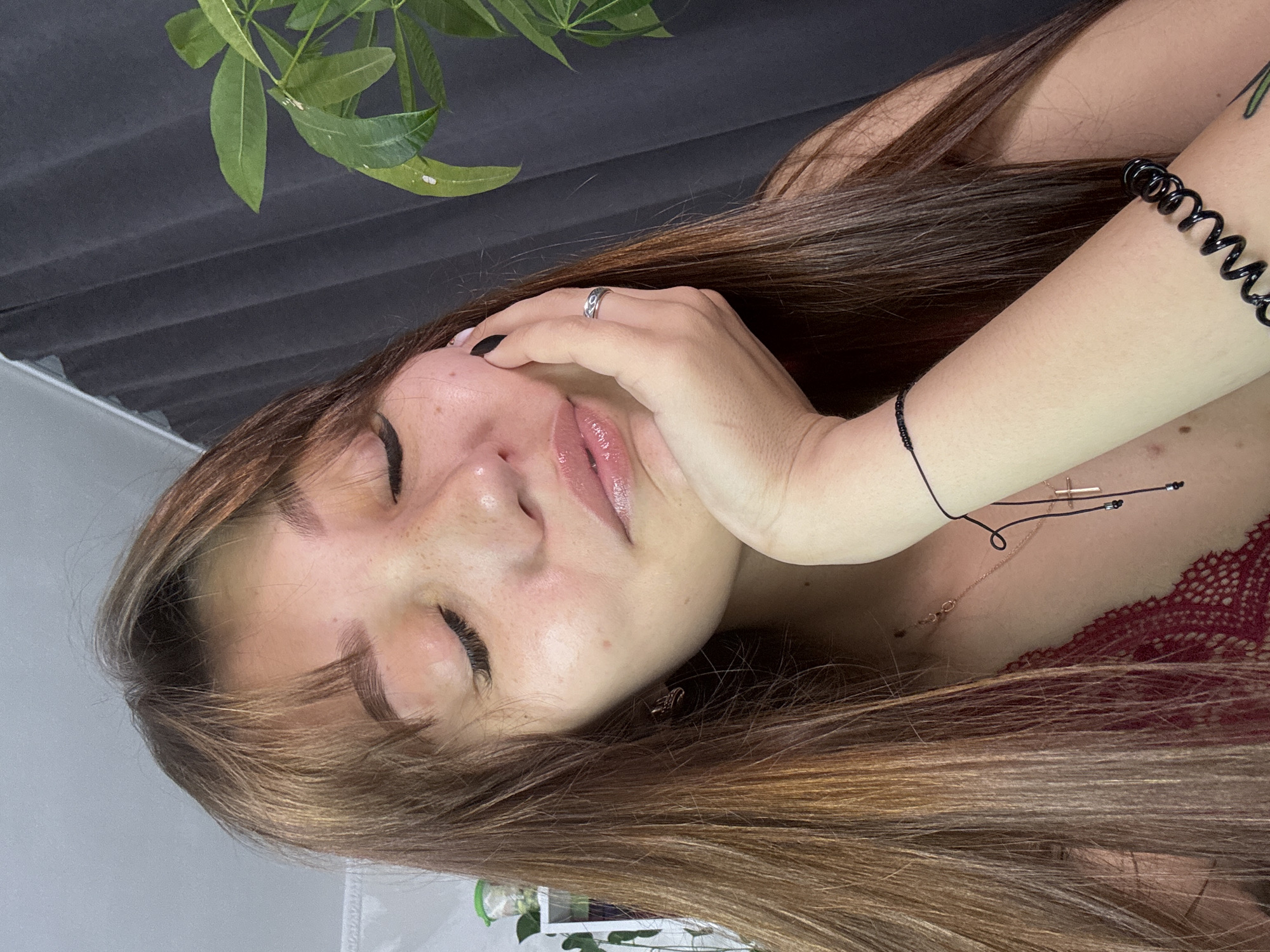 A young woman with long, brown hair and bangs is leaning her head on her hand, with her eyes closed. She is wearing a dark red lace top. She has a ring on her right ring finger and a thin bracelet on her left wrist. A dark gray curtain and a green plant are visible in the background. The image appears to have been taken indoors.