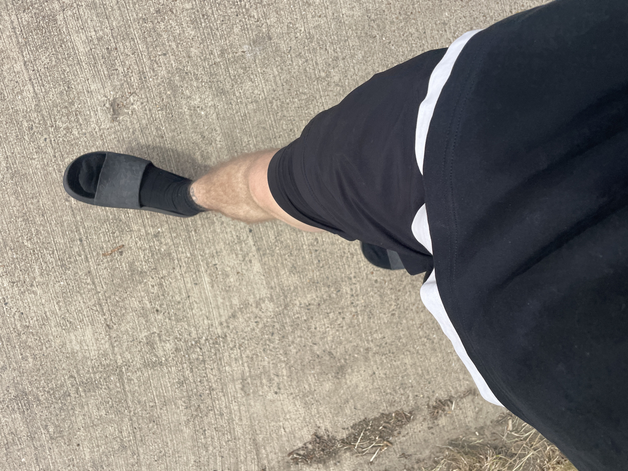 The image shows a person's legs from the thigh down, walking on a concrete surface. The person is wearing black shorts with a white stripe near the hem, black socks, and black slide sandals. The concrete appears to be outdoor pavement, possibly a sidewalk or path, with some dry grass and debris on the left side of the frame. The lighting suggests it is daytime. The image does not provide any specific visual cues that would allow for precise location identification within Diepenbeek, Belgium.