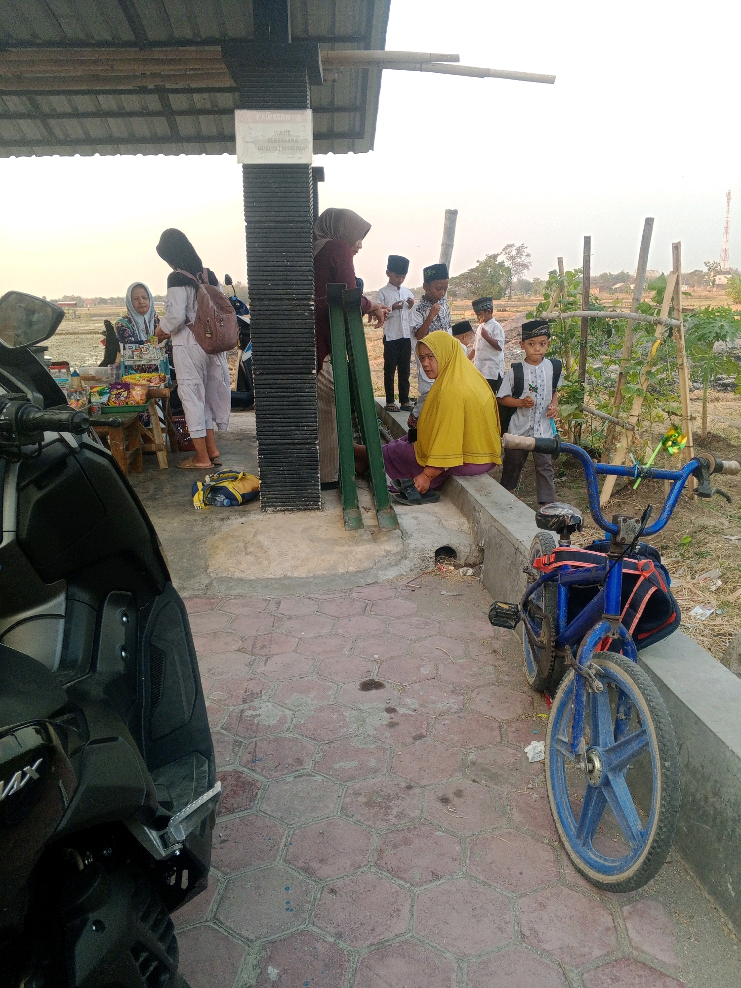 In Lembah, Indonesia, a group of children wearing traditional Muslim attire and peci hats stand near a covered outdoor area with a food stall. A woman in a yellow hijab sits on a concrete ledge, looking down. Another woman, also in a hijab, stands behind a wooden table laden with snacks. A blue bicycle with a backpack attached to the handlebars is parked nearby. In the foreground, a black scooter is partially visible. The background shows a field and a distant telecommunications tower under a hazy sky.