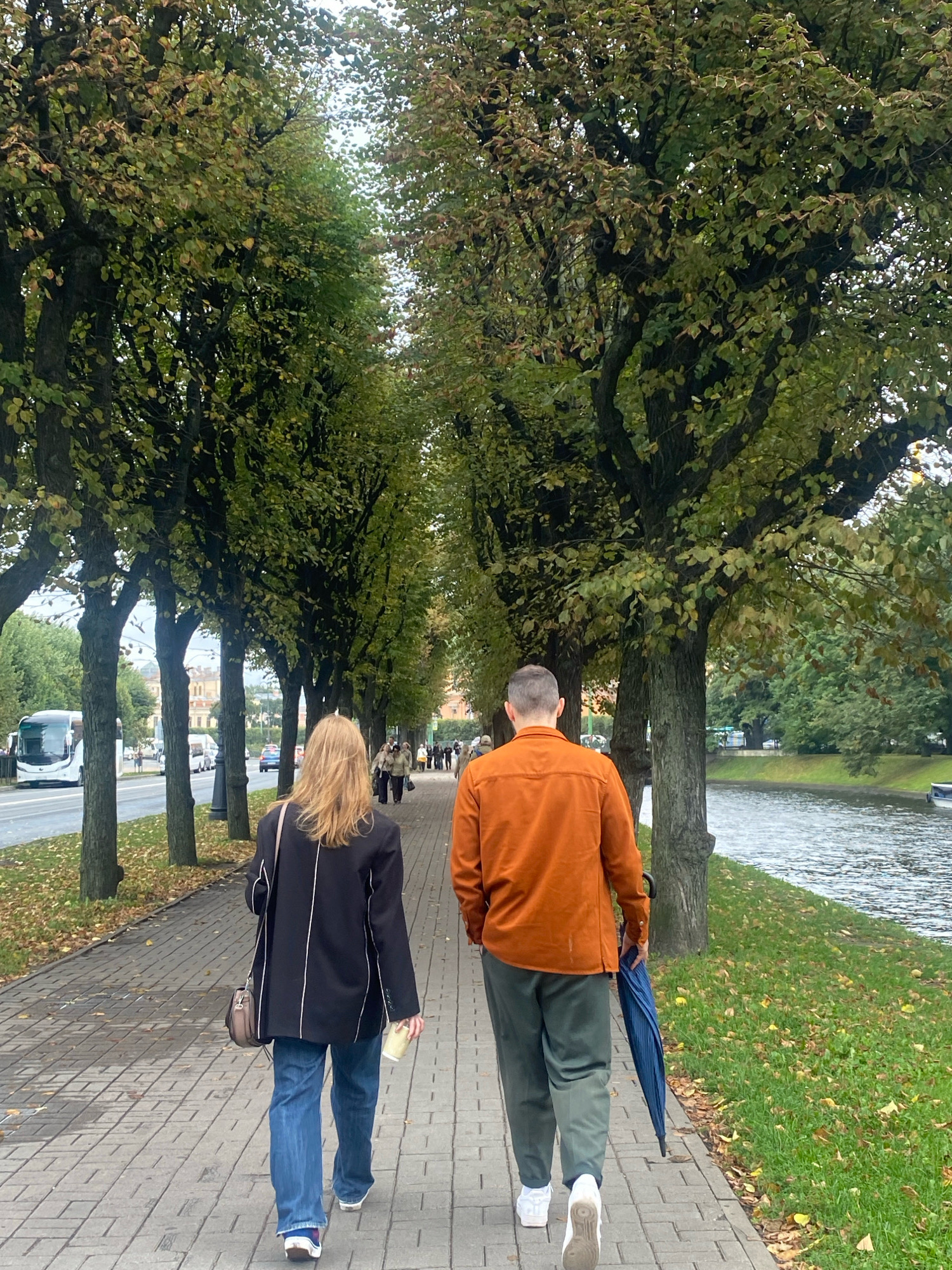 A man and a woman are walking away from the camera on a paved sidewalk lined with trees. The man is wearing an orange shirt and green pants and is carrying a closed blue umbrella. The woman is wearing a black jacket and blue jeans and is holding a cup. The sidewalk is to the left of a street with a bus and cars, and to the right of a canal. The trees have green and yellow leaves, indicating it is autumn. The buildings visible in the background suggest an urban setting, likely in Saint Petersburg, Russia, given the architectural style and the presence of a canal. The overall atmosphere is overcast and cool, typical for a fall day in the city.