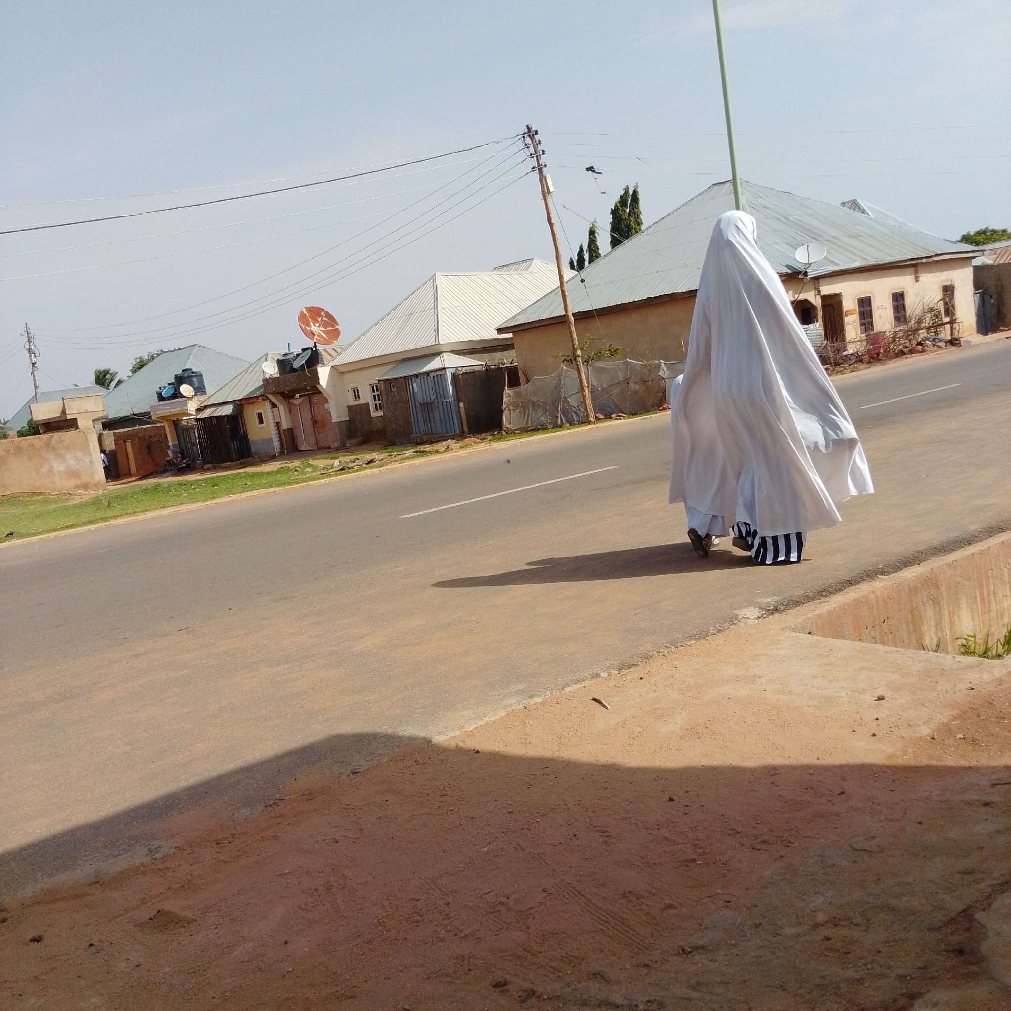 In Gombe, Nigeria, two individuals are walking on a paved road. The person in front is wearing a white garment that covers their entire body and head, creating a voluminous silhouette. They are walking on the right side of the road, facing away from the camera. Behind them, another person, likely a child, is also walking, mostly obscured by the front individual's attire. This person's legs are visible, wearing striped trousers with black and white vertical lines. The background shows a street lined with buildings with corrugated metal roofs, some of which have satellite dishes. Power lines run overhead, supported by wooden poles. The road is asphalt, with a white line marking the center. A sandy embankment with some vegetation is present on the right side of the road. The sky is clear and blue.