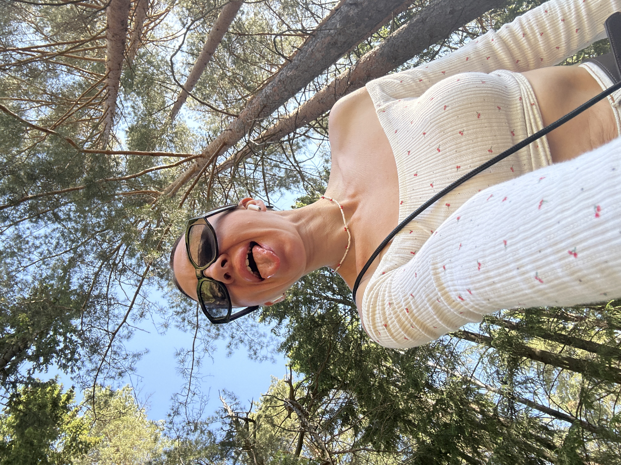 A selfie taken outdoors from a low angle, likely in a wooded area. The subject, a woman, is wearing sunglasses, an earbud in her ear, a beaded necklace, and a white long-sleeved shirt with small red details. She is sticking her tongue out. Overhead are tree branches and sky. The location cannot be determined to be in Votkinsk, Russia, solely from the given image.