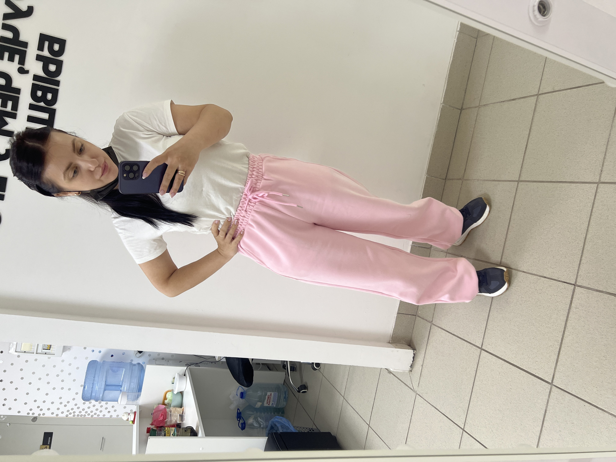 The image displays a mirror selfie, rotated 90 degrees clockwise, featuring a woman standing indoors. She has long, dark hair, pulled back, and is wearing a white short-sleeved t-shirt and light pink sweatpants with a drawstring waist. She completes her casual outfit with dark blue sneakers that have light brown soles. In her right hand, she holds a dark-colored smartphone, likely an iPhone given its camera array, using it to take the picture. A gold-colored ring is visible on her right ring finger, and her left hand rests on her hip. Her expression is neutral, focused on the phone screen.
The setting is a room with bright white walls and light grey tiled flooring. In the background, visible in the mirror's reflection, there is large black text on the white wall, appearing upside down and mirrored. It seems to read "MADE IN" followed by a partially obscured word like "EPISODE" or "DEPO". Below this text, a counter or desk area holds a large blue water dispenser bottle, alongside several smaller plastic bottles. Faint Cyrillic text, possibly "ЛИТВИНОВ АРХОТОМ", can be seen on one of the water bottles. The scene suggests a casual indoor environment, possibly an office, gym, or retail space.