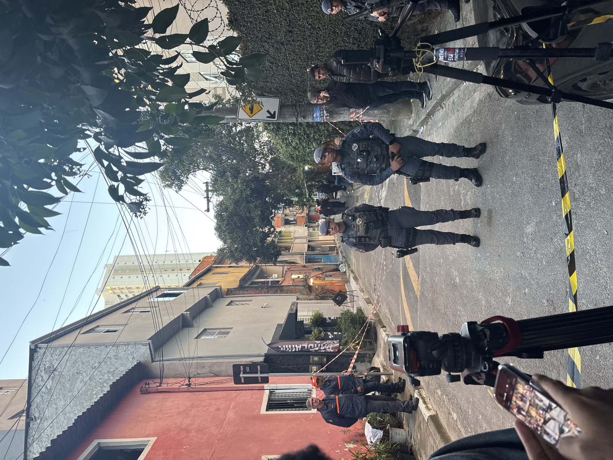 The image depicts an urban street scene in São Paulo, Brazil, under clear daytime skies. The street, paved with asphalt and marked with yellow lines, appears partially cordoned off. In the immediate foreground, a person's hand holds a smartphone, partially obscuring a black camera rig or crane. This rig is wrapped with yellow and black caution tape, on which the Portuguese text "EU ARTICULA" is visible.
Two uniformed police officers, dressed in blue tactical gear with vests and caps, stand in the middle of the street, seemingly monitoring the area. Further back, two other individuals in dark civilian clothes are present near additional camera equipment. Towards the lower left, two men wearing orange and blue work uniforms are visible near a red-colored building.
The street is lined with various buildings of diverse architectural styles and colors, including red, grey, and yellow. One building features a corrugated metal facade. A black banner hanging from a building on the left side displays the word "PARCERIA" in white text. Utility poles crisscrossed with numerous overhead wires are prominent against the sky. A wall topped with barbed wire is visible in the upper left. A yellow road sign with a black arrow indicating a sharp turn or curve is attached to a pole.
The presence of multiple uniformed personnel, various camera setups, and caution tape suggests a controlled environment, possibly related to a security operation, a media production, or an ongoing incident response. The overall atmosphere appears orderly and calm, with no overt signs of distress.