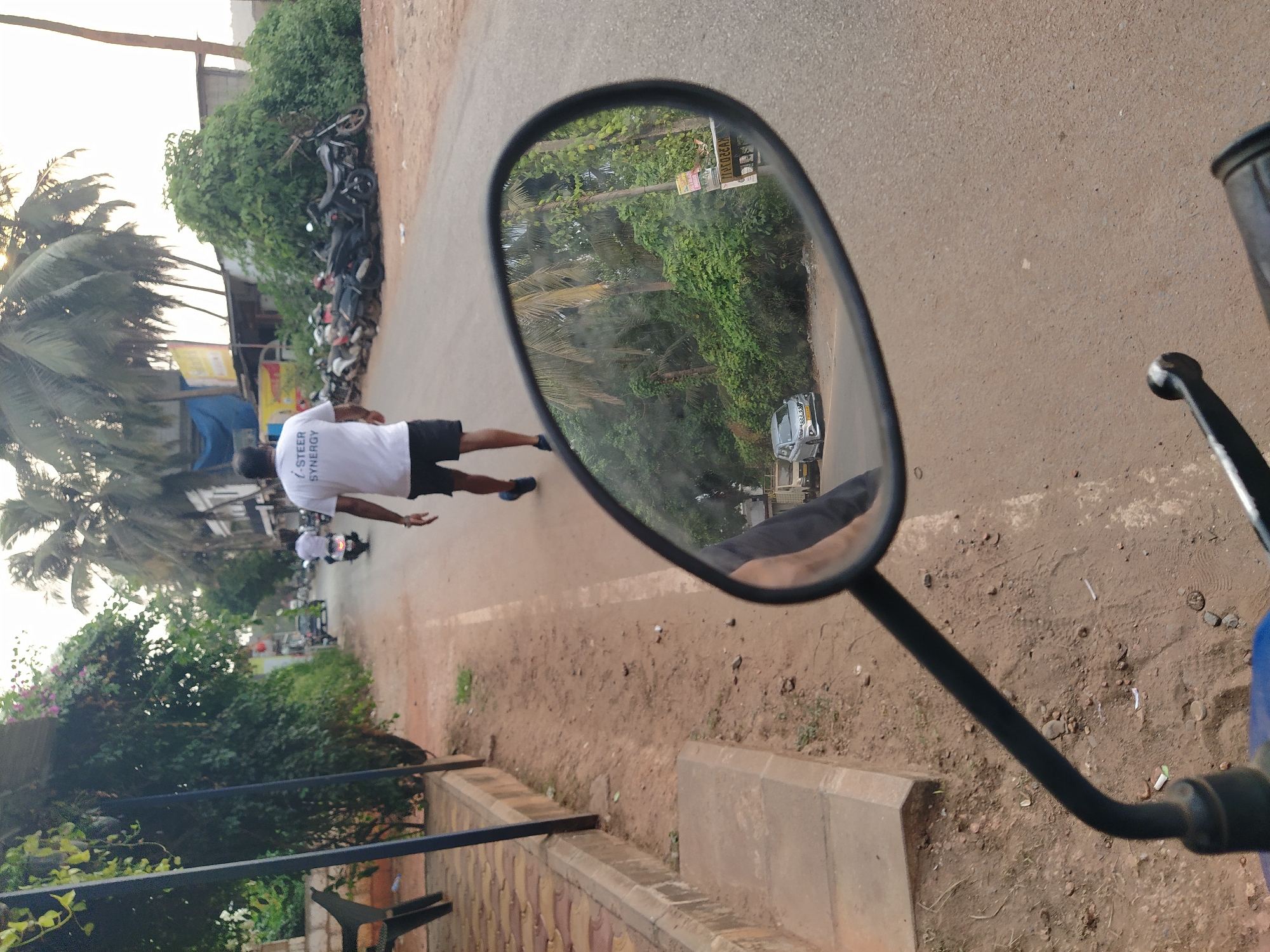 The photograph depicts a street scene in Oxel, India, viewed through the side mirror of a motorcycle. In the reflection, a man wearing a white t-shirt and black shorts is walking along the road. A scooter follows him, and several motorcycles are parked on the side of the road. Lush green foliage, including palm trees, lines the street. A car is also visible in the mirror's reflection. The foreground shows the motorcycle's handlebars and the surrounding ground, which is dirt and gravel with sparse vegetation. A brick curb is also visible in the foreground.