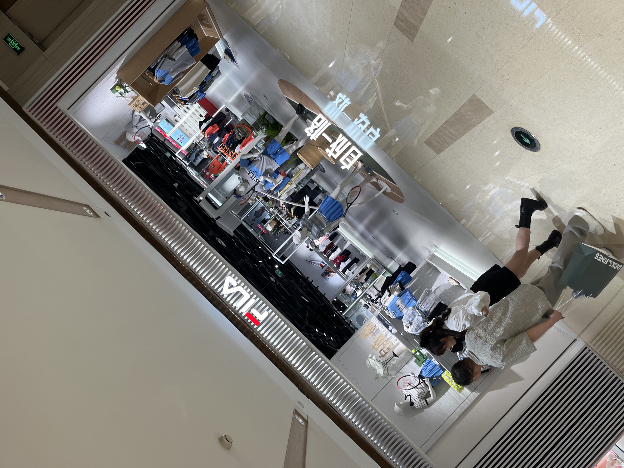 The image shows the interior of a Fila store in a shopping mall, likely located in the Yuzhong District of Chongqing, China. The store has a modern design, with mannequins displaying athletic apparel. The Fila logo is prominently displayed on the wall. There are several rows of clothing racks. Two people, a man and a woman, are standing by the storefront, looking at the merchandise. The woman wears a black skirt and a white top. The man is wearing a gray shirt. They have shopping bags from JACK&JONES. The floor of the mall is light colored.