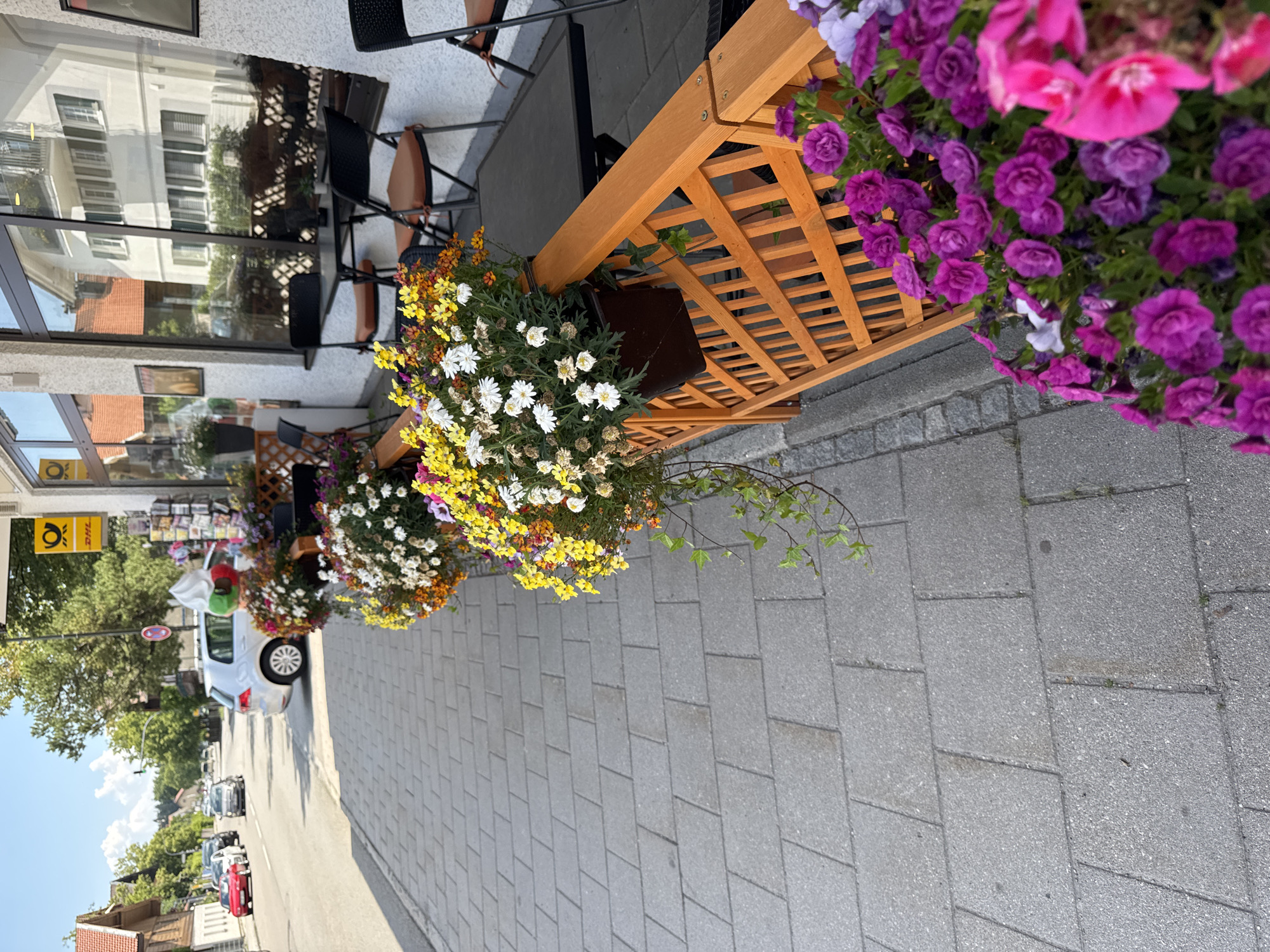 The image shows a street scene in Höhenkirchen-Siegertsbrunn, Germany, likely on a sunny day. The foreground features a sidewalk paved with gray rectangular tiles. Along the edge of the sidewalk, a wooden planter box with a lattice design holds an abundance of colorful flowers, including purple, pink, and white varieties. Behind the planter, a dark-colored table and chairs are set up, suggesting an outdoor seating area for a business, possibly a cafe or shop.
Further into the image, a white car is parked on the street. A building with windows and architectural details is visible to the right, reflecting the sky and surrounding environment. On the left side of the street, more buildings and trees can be seen. The street itself has other vehicles, including a red car and a silver car further down, indicating it is a moderately busy road. A yellow and blue DHL/Post sign is mounted on the building to the right. The overall scene is characteristic of a European town, with a focus on pedestrian-friendly spaces and decorative elements.