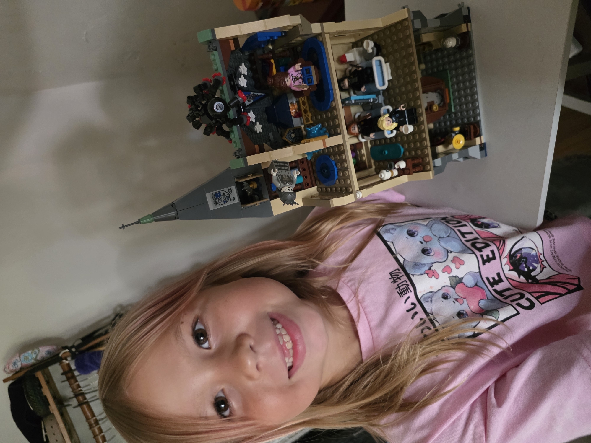 A young girl with blonde hair, light pink highlights, and a bright smile is positioned in the foreground. She is wearing a pink t-shirt with a graphic design featuring cartoon animals and Japanese text. Behind her is a detailed Lego set of a building, featuring multiple floors and various rooms filled with Lego figures and accessories. The building appears to be displayed on a white surface, likely a table, and the background reveals a wall with hanging items. The location, North Kuta, Indonesia, is not evident in the image.
