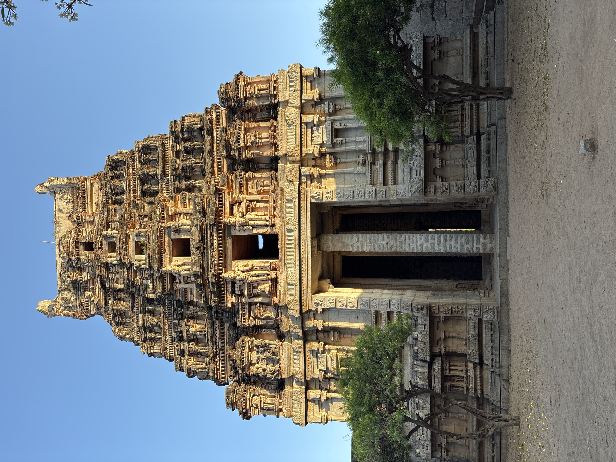 The image captures the Hazara Rama Temple in Kamalapura, India. The temple's towering gopuram, adorned with intricate carvings, dominates the frame. The temple entrance is flanked by carved pillars and stone walls. Two trees with green foliage frame the lower part of the structure. The ground is visible, a tan color. The sky above the temple is a clear blue.