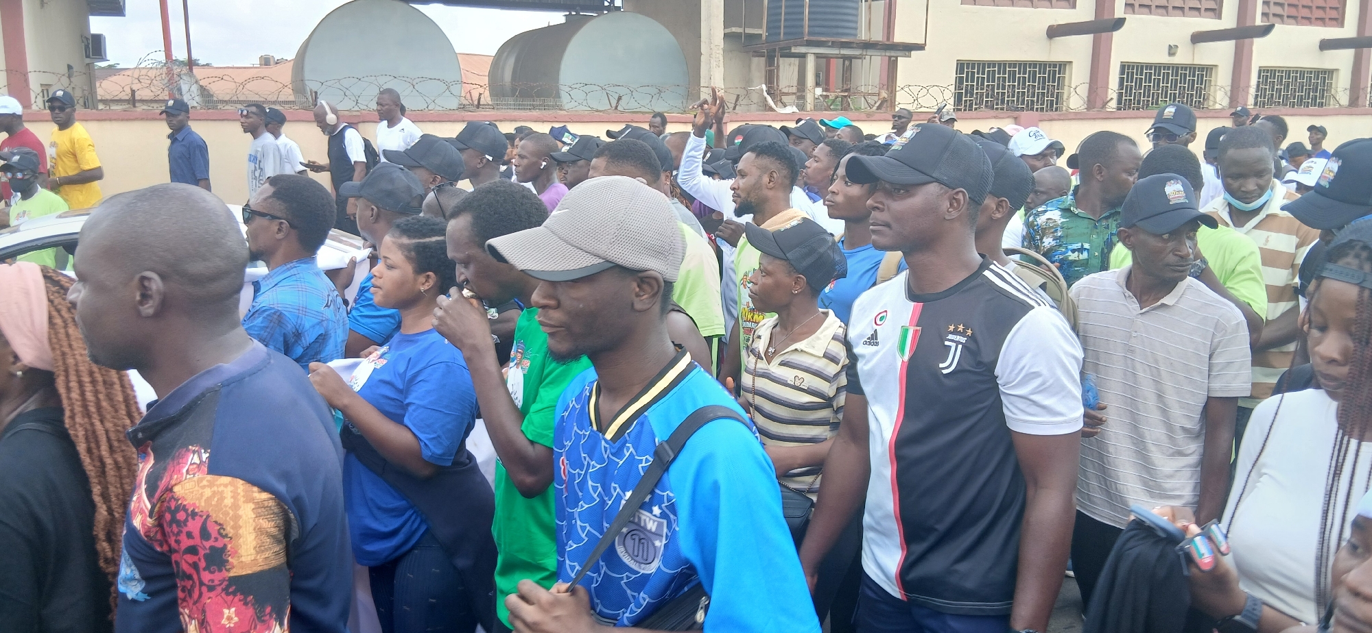 The image depicts a crowd of approximately 30 to 40 individuals, predominantly men, gathered outdoors in what appears to be an urban setting. The background includes a building with a textured facade, possibly housing industrial tanks or machinery, and a fence topped with barbed wire. The ground surface is not clearly visible, but the overall impression is one of a public space or gathering area.
The individuals are dressed in casual attire, with a variety of t-shirts, shirts, and caps visible. Several people are wearing face caps. Some individuals are holding items, including a phone being held by a woman in the foreground, and what appears to be paper or leaflets being held by some in the middle ground. The people are facing in various directions, suggesting they are engaged in conversation or observing something of interest. The crowd appears to be in motion or anticipating movement, as some are looking forward and others are in profile.
The lighting suggests it is daytime, possibly overcast. The image is taken from a slightly elevated perspective, looking across the crowd. The specific location within the city of Tudun Nupawa, Nigeria, is not directly indicated by any visual cues in the image itself.