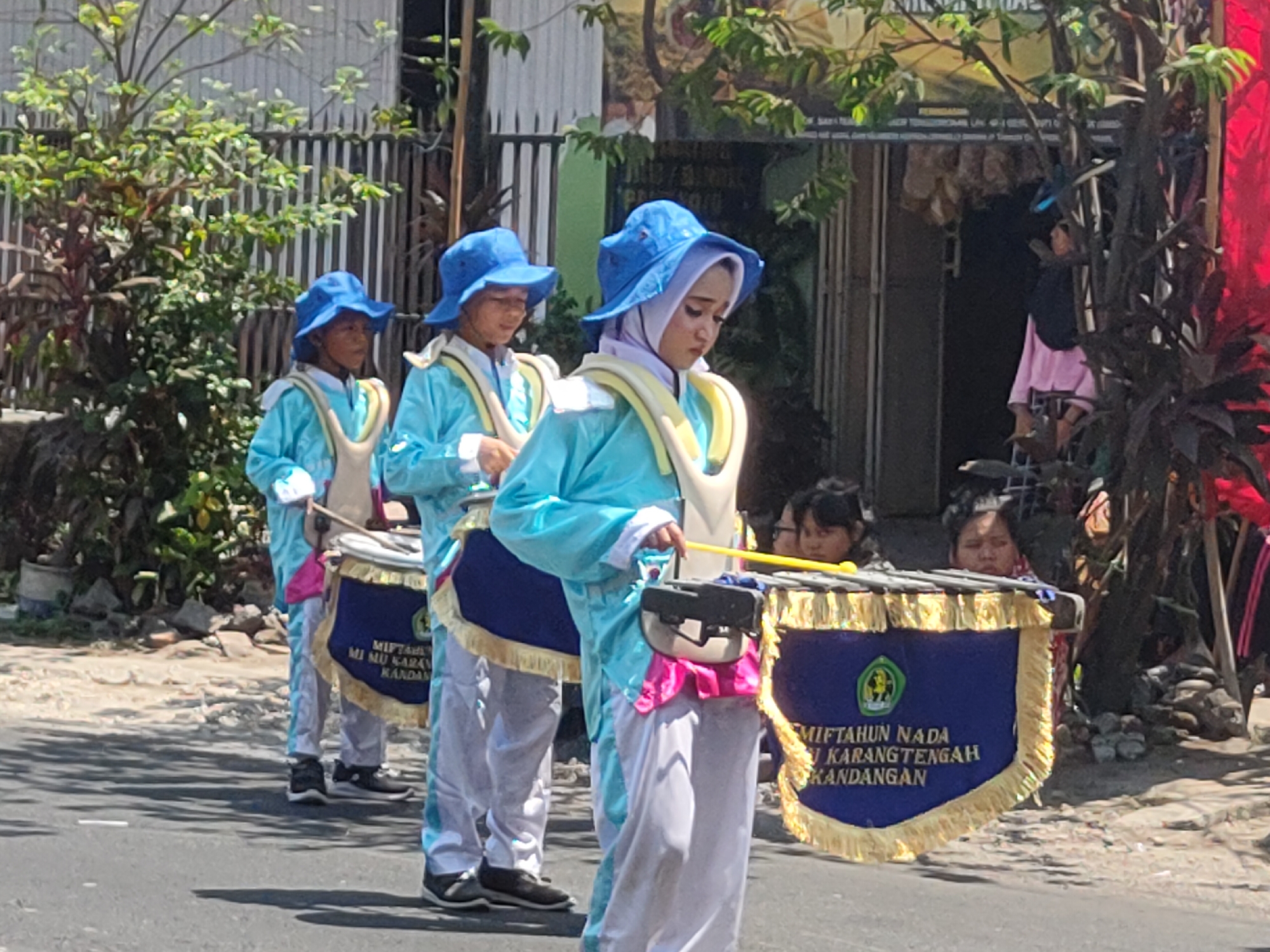 In Ngasem, Indonesia, three young individuals are participating in a parade. They are dressed in matching bright turquoise uniforms with white accents and blue hats. Each individual is carrying a drum, with the closest person to the foreground holding a drum decorated with a blue banner featuring yellow fringe and a green emblem with text that reads "MIFTAHUN NADA MI MU KARANGTENGAH KANDANGAN." They are positioned on a paved road, with onlookers and buildings visible in the background. The scene suggests a celebratory or cultural event.