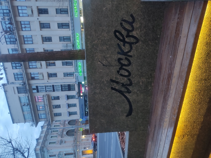 The image depicts a view of Moscow, Russia. A building with multiple windows and the name "GLENFIELD" visible is in the background. In the foreground, there is a structure with the word "Москва" (Moscow) written on it in a cursive style. The structure appears to be part of a seating area with wooden planks and embedded lighting. A street with a car is also visible. The sky is overcast.