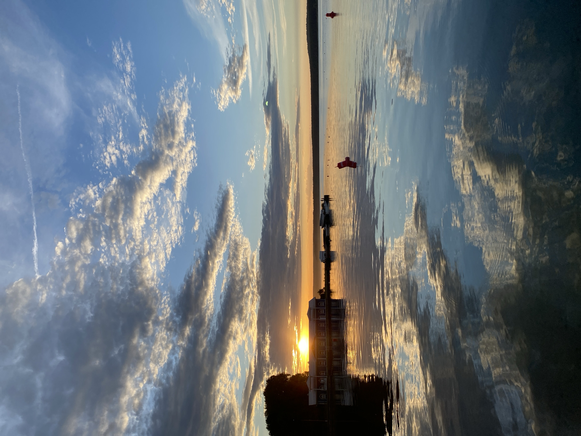 This is a vertically oriented, low-angle photograph of a sunset over a body of water, likely a lake or river. The sky is a mix of blue and white, with dramatic clouds that are illuminated by the setting sun, creating a warm glow. The sun itself is a bright, almost blinding orb peeking through the clouds just above a building.
The main subjects in the scene are the sky, the water, and a long, multi-story building reflected in the water. The building appears to be a hotel or some sort of residential complex, with multiple windows visible. In front of the building, there are trees silhouetted against the light. In the distance, along the horizon line, there is a dark, forested shoreline. On the water, there are several red buoys, some with white markings that could be text, though it is not legible. There are also some small boats or docks visible near the building.
The reflection of the sky, clouds, and building in the calm water is a prominent feature, almost mirroring the scene above. The water's surface is disturbed by ripples, creating abstract patterns of light and color. The overall mood is serene and peaceful, with a sense of natural beauty enhanced by the warm colors of the sunset. The time of day is clearly sunset, given the low angle of the sun and the warm lighting. The weather appears clear with scattered clouds. Given the location context of Votkinsk, Russia, this is likely a scene on the Kama River or a nearby reservoir. There is no discernible text in the image.