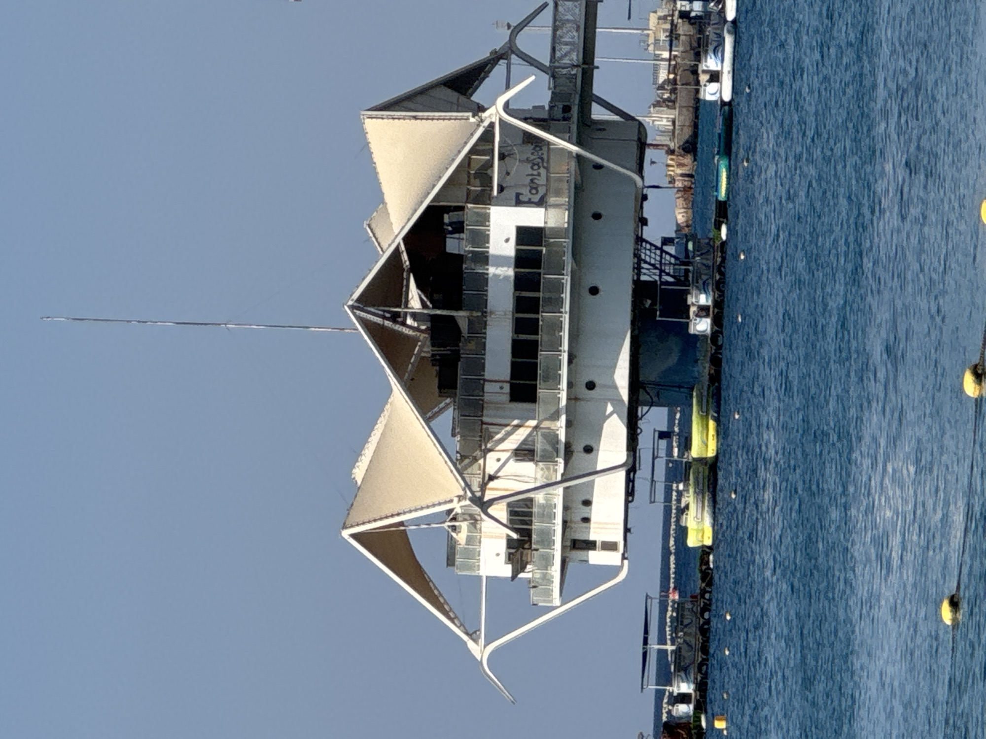 The image shows a floating structure labeled "Fantasea" in the city of Eilat, Israel. The structure is white with a large, light-colored canopy supported by metal beams. It has multiple levels with windows and railings. The structure is situated in blue water, and several smaller boats and buoys are visible in the foreground and surrounding water. In the background, a tall antenna or mast rises from the structure, and a hazy cityscape or shoreline can be seen. The sky is clear and blue. The scene appears to be a sunny day.