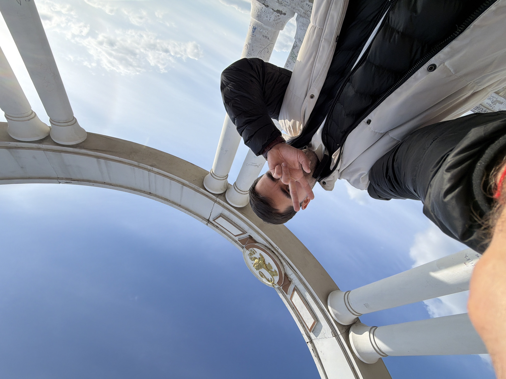 The image shows a person standing on a bridge-like structure in Krasnoyarsk, Russia, taking a selfie. The structure is an archway with white pillars and a decorative emblem in the center. The sky is visible in the background, transitioning from a clear blue to scattered clouds. The person is wearing a winter coat and holding their hand up to their face.