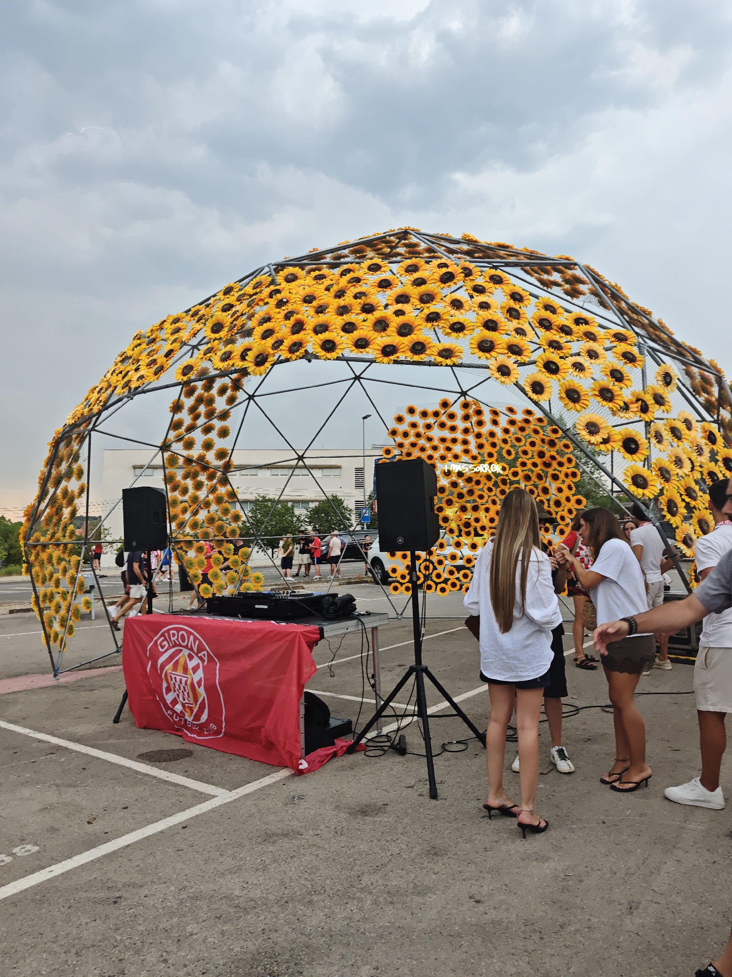 An outdoor event is taking place in a parking lot. The main subject is a large geodesic dome structure adorned with numerous artificial sunflowers. In front of the dome, a red tarp with a crest is spread over a table that holds DJ equipment and speakers. Several people are gathered around the dome and DJ setup. Some are standing and conversing, while others appear to be observing the event. The ground is paved, and white lines mark parking spaces. The sky is overcast with clouds.