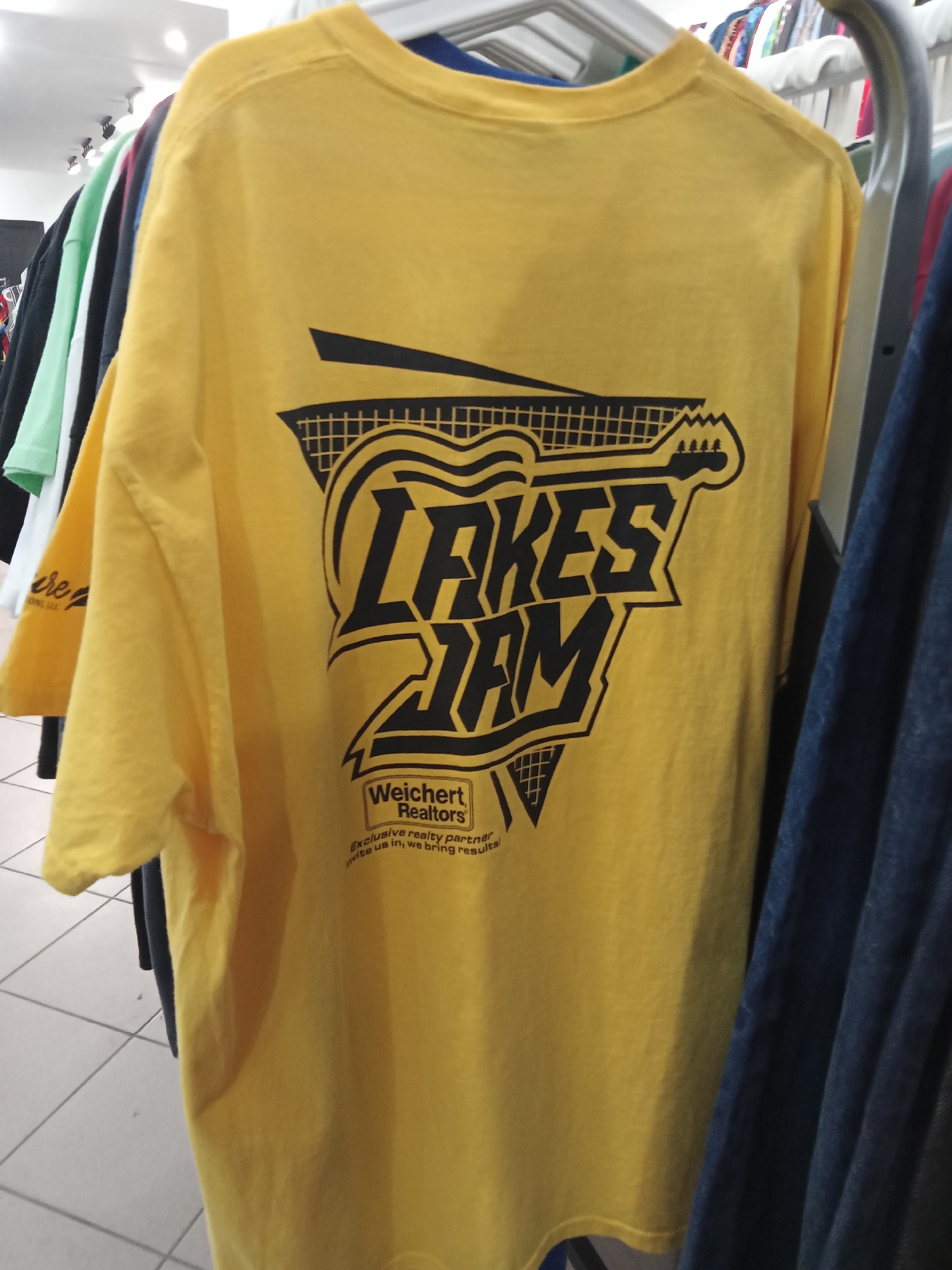 A yellow t-shirt with a black graphic design of a guitar and the text "LAKES JAM" is displayed on a clothes rack in a store. Below the "LAKES JAM" text, the words "Weichert Realtors" are visible, along with smaller text that reads "Exclusive realty partner invite us in, we bring results." The t-shirt is hanging with other shirts in various colors, including green, black, and white, on a metal rack. The background shows a tiled floor and clothing racks in a retail environment.