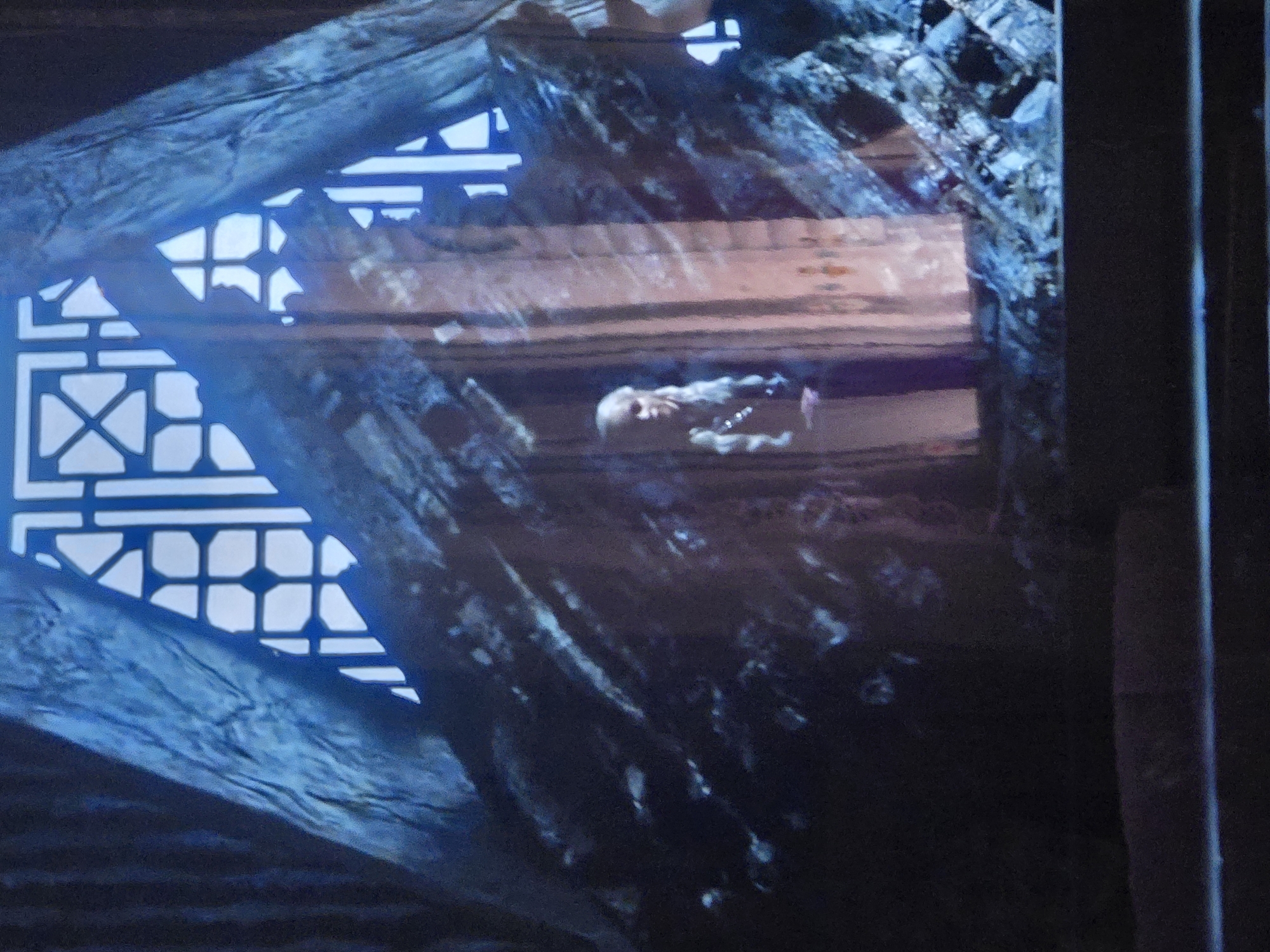 In a dimly lit, cavernous interior, a woman with long, blonde hair braided in multiple strands stands behind a rocky, crystalline structure that is illuminated by a warm, orange light. She wears a dark, textured garment. Behind her, a large, angular structure with geometric cutouts, possibly a window or a screen, is brightly lit with white light. The foreground is dominated by dark, rocky debris. The scene appears to be a fantasy or historical setting, possibly a throne room or a sacred space, within the city of Unknown, Unknown.