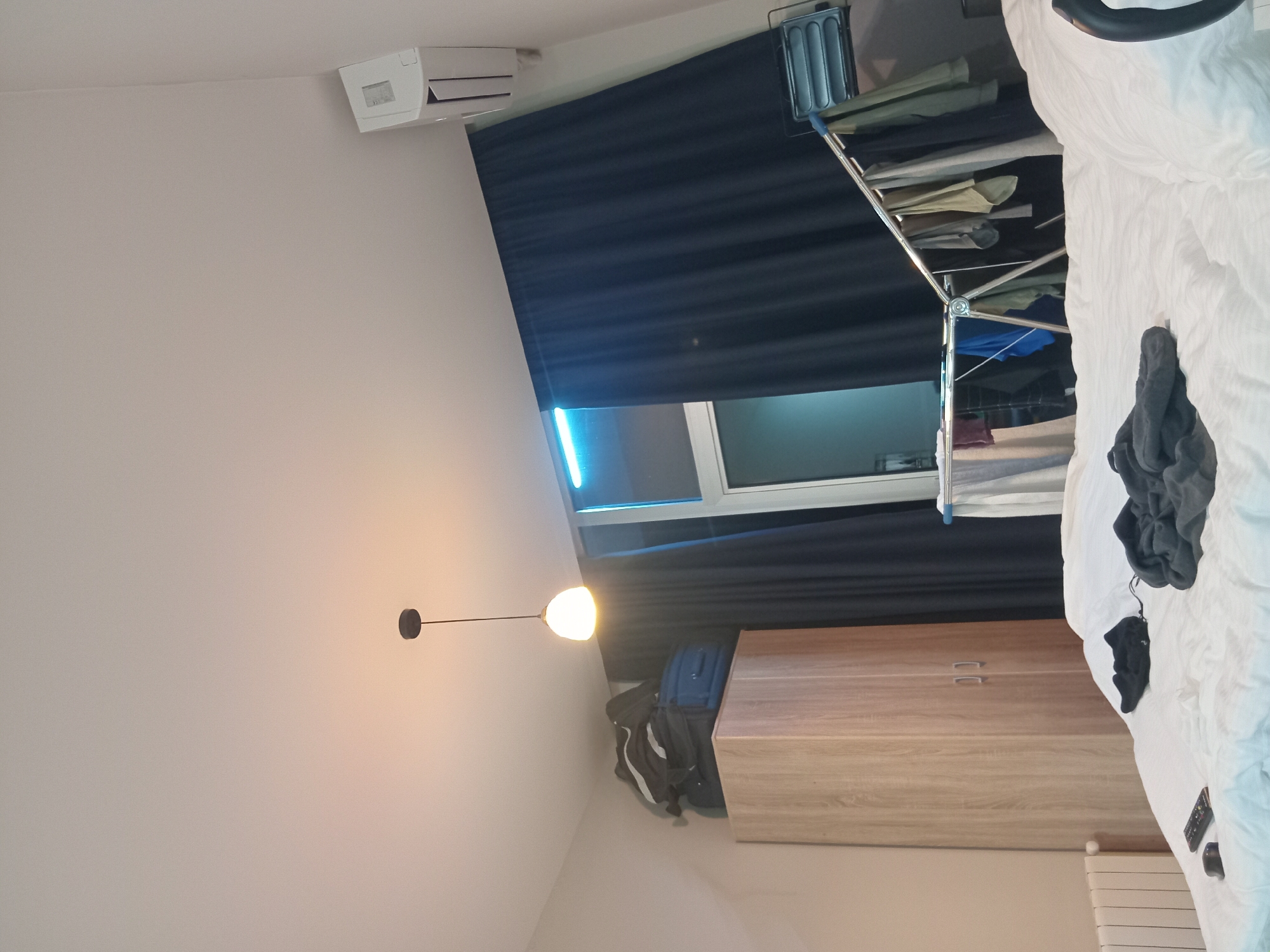 This is an interior shot of a room, possibly in a residence. The photo contains a bed with white linens, a folding clothes drying rack with various items of clothing, a cabinet with two drawers, a lamp, an air conditioning unit, and luggage. The room has a window with black curtains. It's difficult to pinpoint the exact location in Batumi, Georgia without more contextual clues. There are no people visible in the frame.