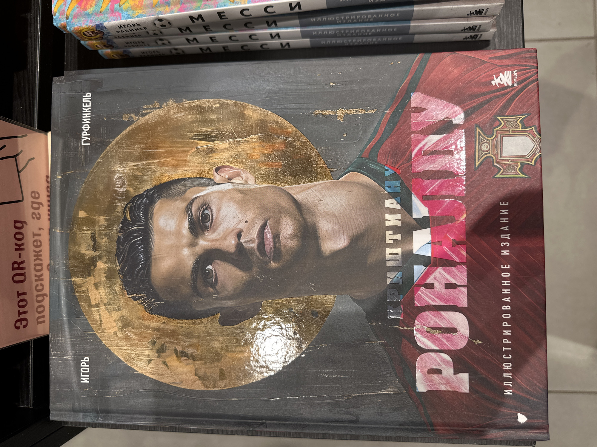 The image shows a book cover titled "Cristiano Ronaldo", subtitled "Illustrated Edition". The book is written by Igor Gurfinkel, and is published by Bombora. The cover features a portrait of Cristiano Ronaldo with a gold halo-like circle behind his head. There are three books on top of this one which are "Messi" illustrated version. The books are most likely inside a store.