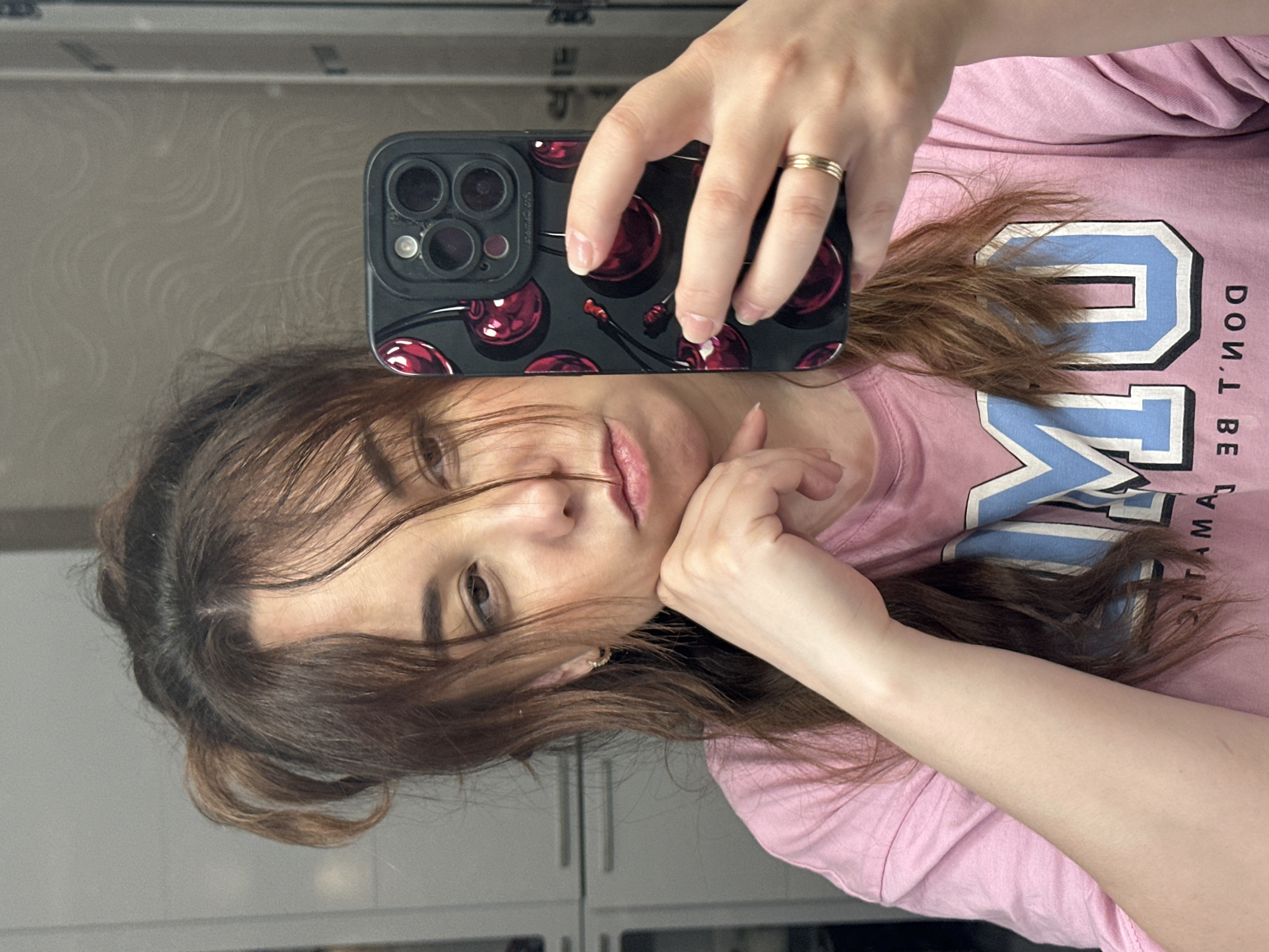 A selfie of a fair-skinned woman with brown hair. She is wearing a pink t-shirt with a blue and white graphic print. The woman is holding up an iPhone with a black case decorated with red cherries. She has a gold ring on her finger. The background appears to be a room with white cabinets. The location is in the city of Unknown, Unknown.